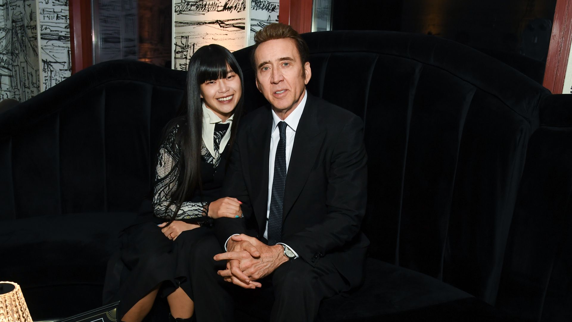 A man in a suit and a woman in a lace dress sit close together on a dark velvet couch in a chic lounge, smiling. A glass coffee table with a lamp and menus sits in front.