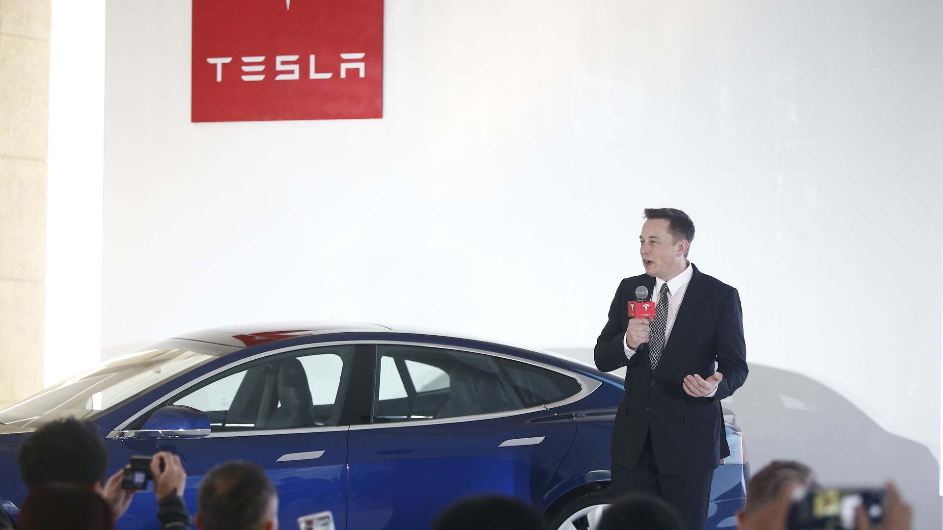Tesla CEO Elon Musk speaking to a crowd in front of a Tesla car.