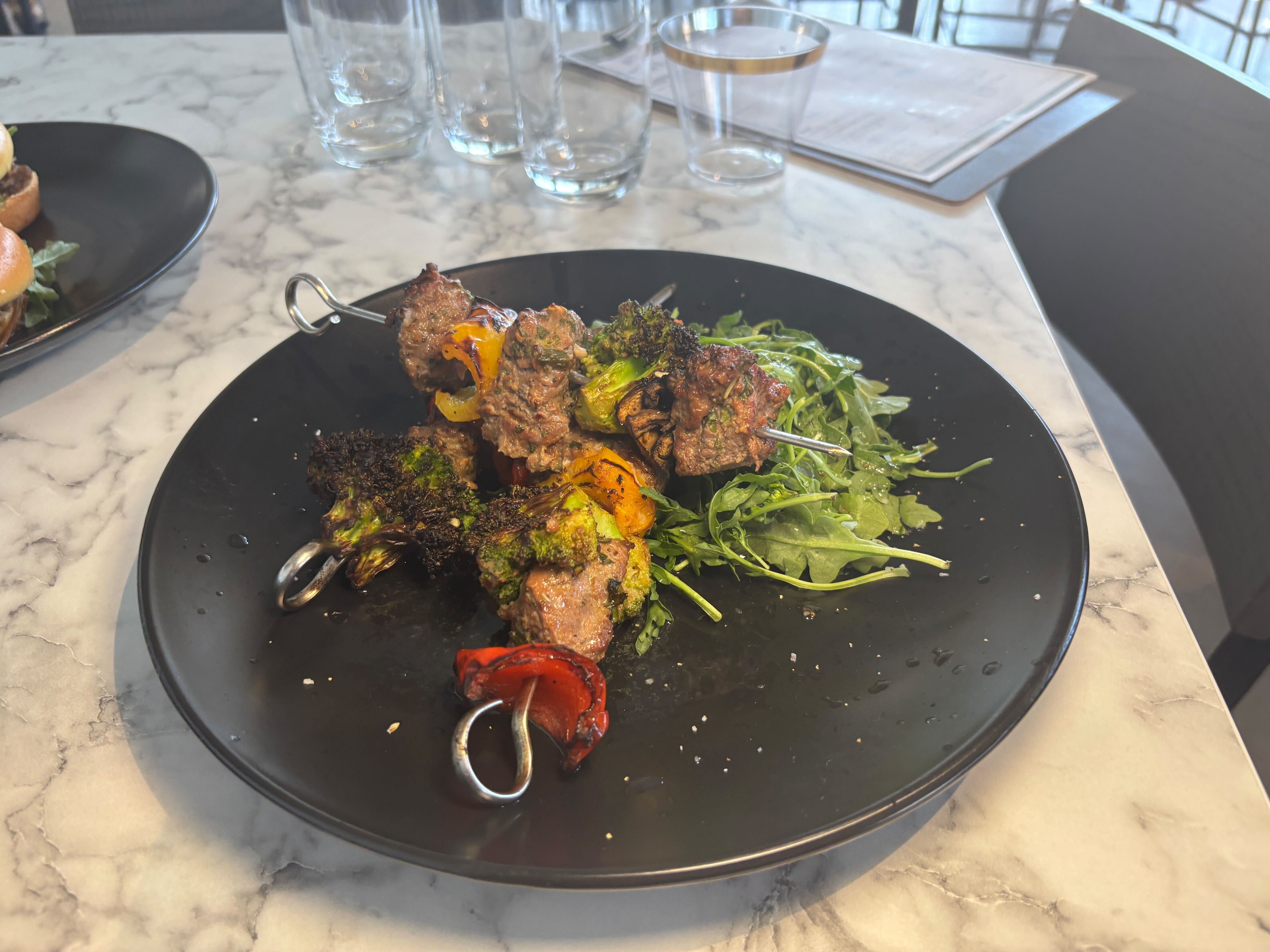 Grilled meat and vegetable skewers with charred broccoli, bell peppers, and arugula on a black plate placed on a white marble table in a restaurant setting.