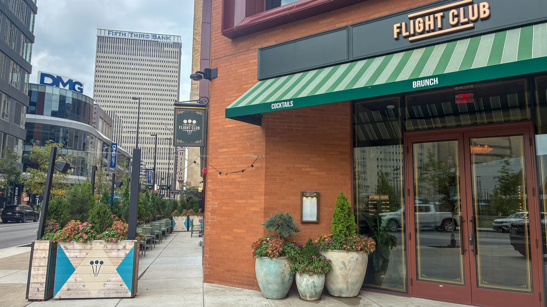 City street with a brick building marked Flight Club under a green striped awning reading COCKTAILS and BRUNCH. Outdoor seating, large planters, glass storefronts; tall office towers and a DMG sign.