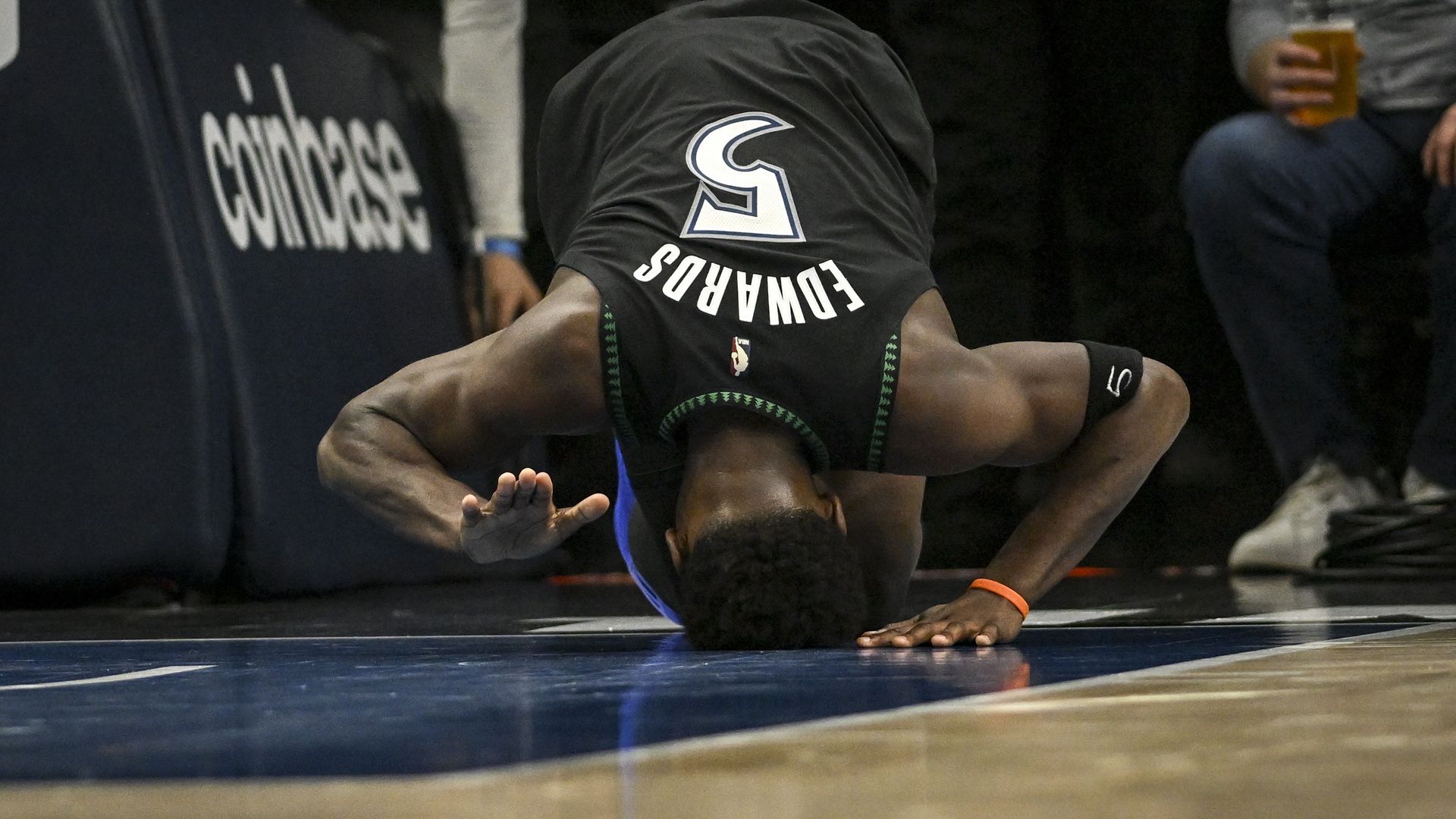 A basketball player in a black jersey tumbles on the court, back to the camera. The jersey features a large white S logo, number 9, and the word FORWARDS with green trim. Spectators in a dark background watch.