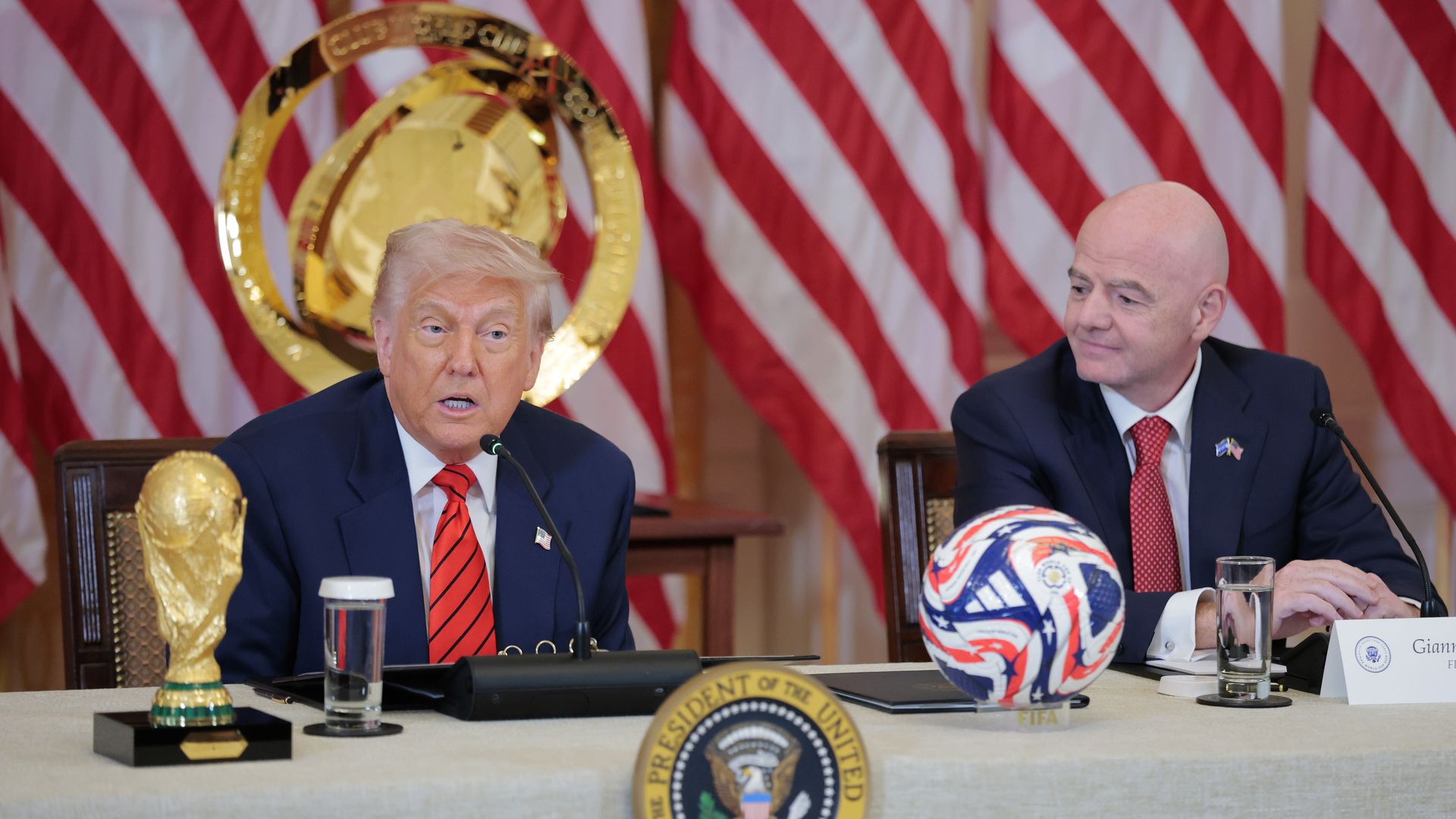 President Trump, wearing a navy jacket with a US flag pin at the top of his left lapel, white shirt and red tie with black stripes, speaks alongside FIFA president Gianni Infantino, wearing a navy jacket, white shirt and red tie with white dots, at a White House desk on which sits the gold World Cup