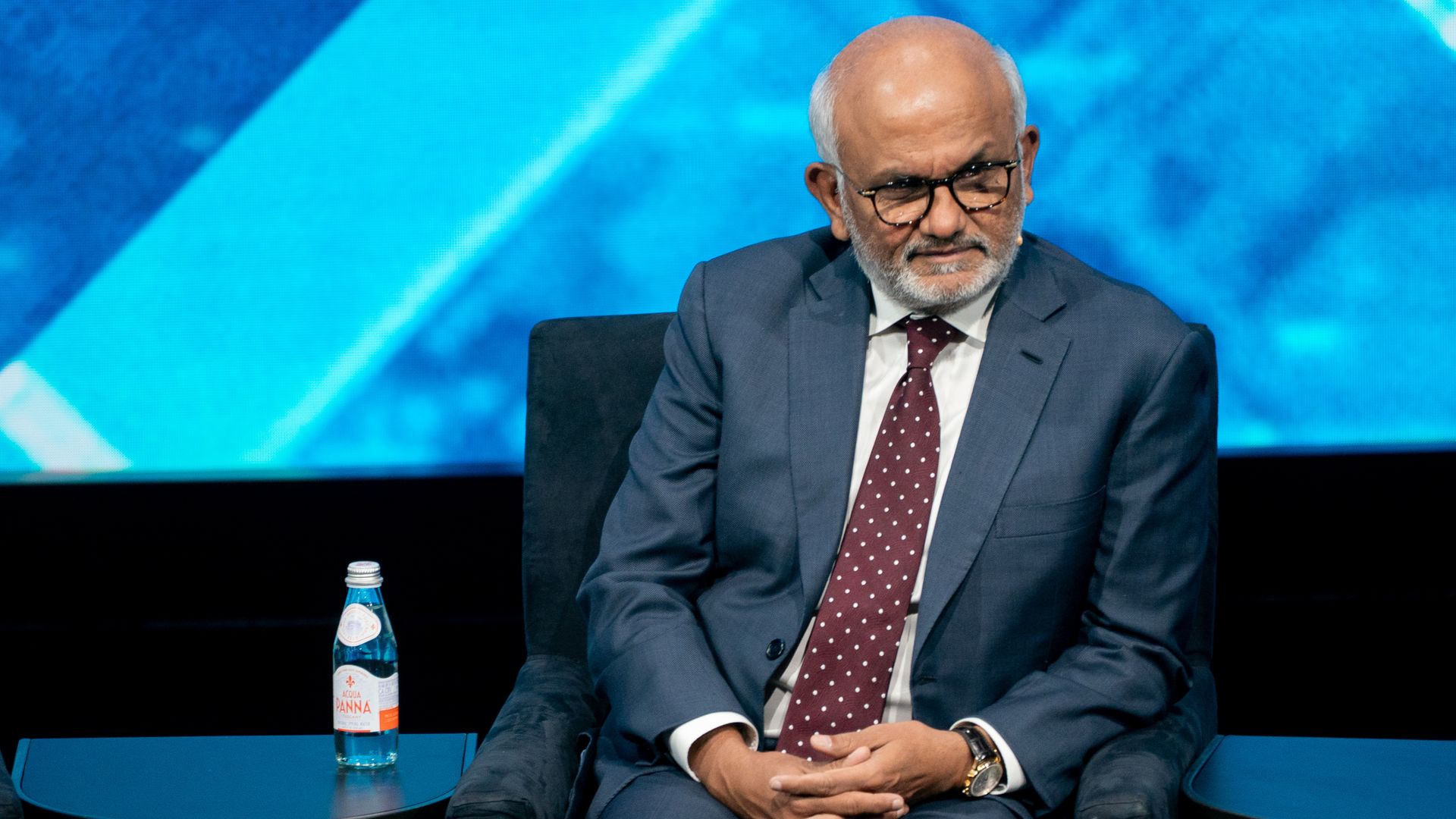 A person n a blue suit and maroon polka-dot tie sits on stage in a dark chair, wearing glasses and a watch. A bottle of water sits on a small table beside him against a blue backdrop.