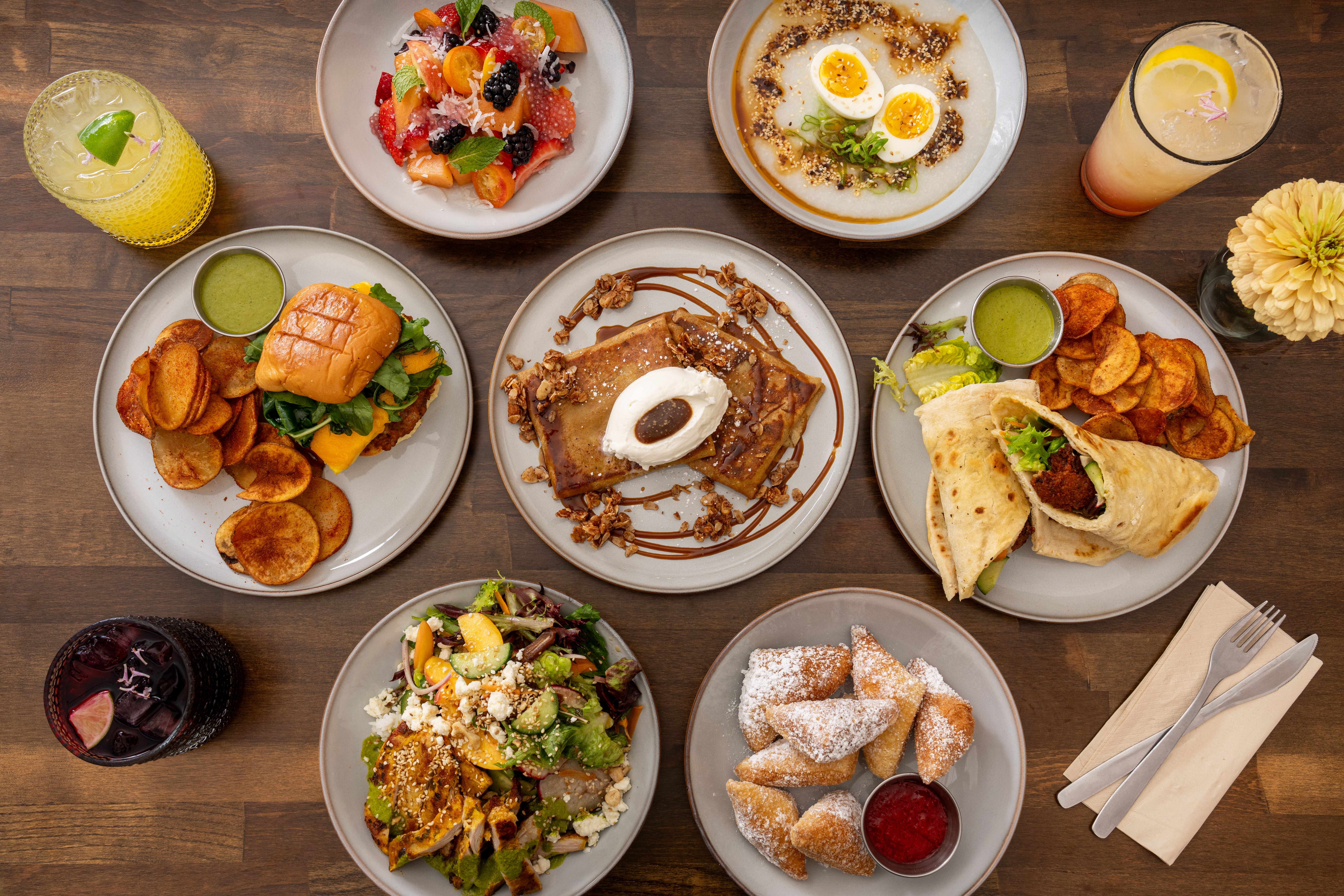 Top-down view of a wooden table filled with colorful dishes: fruit salad, egg bowls, a burger with chips and green sauce, a burrito, pancake with granola, a salad, two citrus drinks, and a pale yellow flower vase.