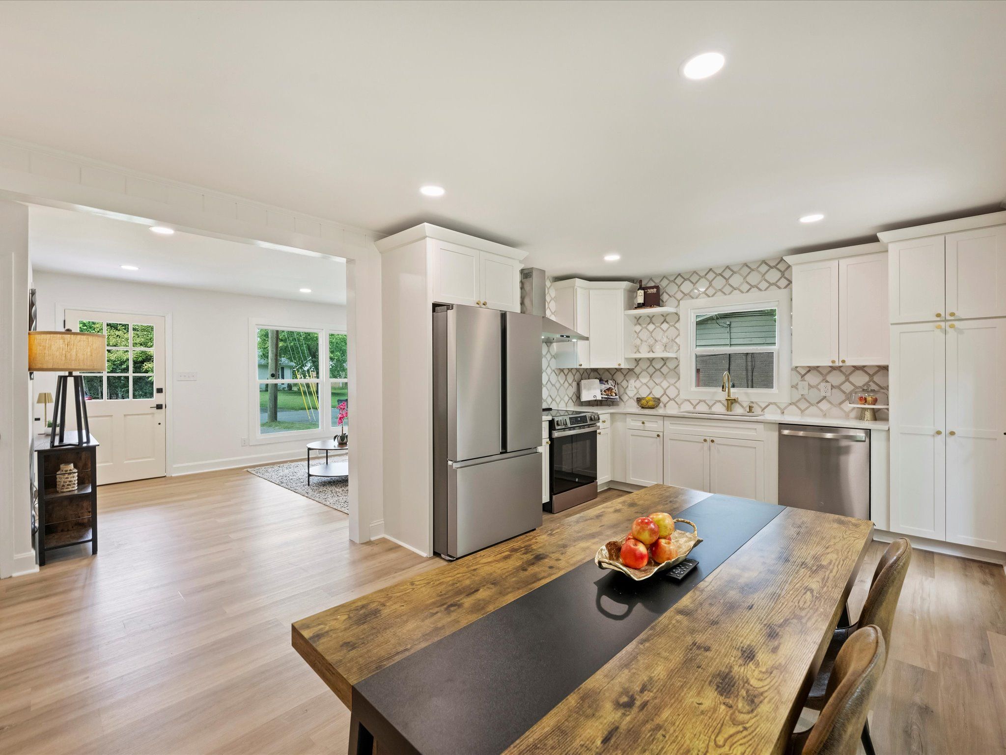 Modern kitchen with white cabinets, stainless steel appliances, patterned tile backsplash, and a wooden table with apples in a golden bowl, adjacent to a bright living room area.