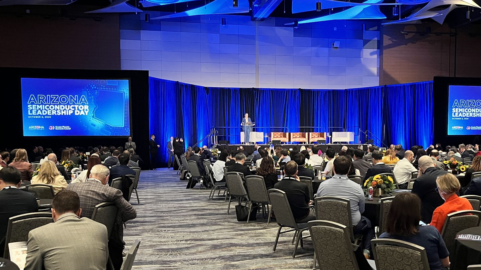 Large conference room with many attendees seated at round tables, a speaker at a podium on stage, and blue screens displaying "Arizona Semiconductor Leadership Day, October 6, 2023".