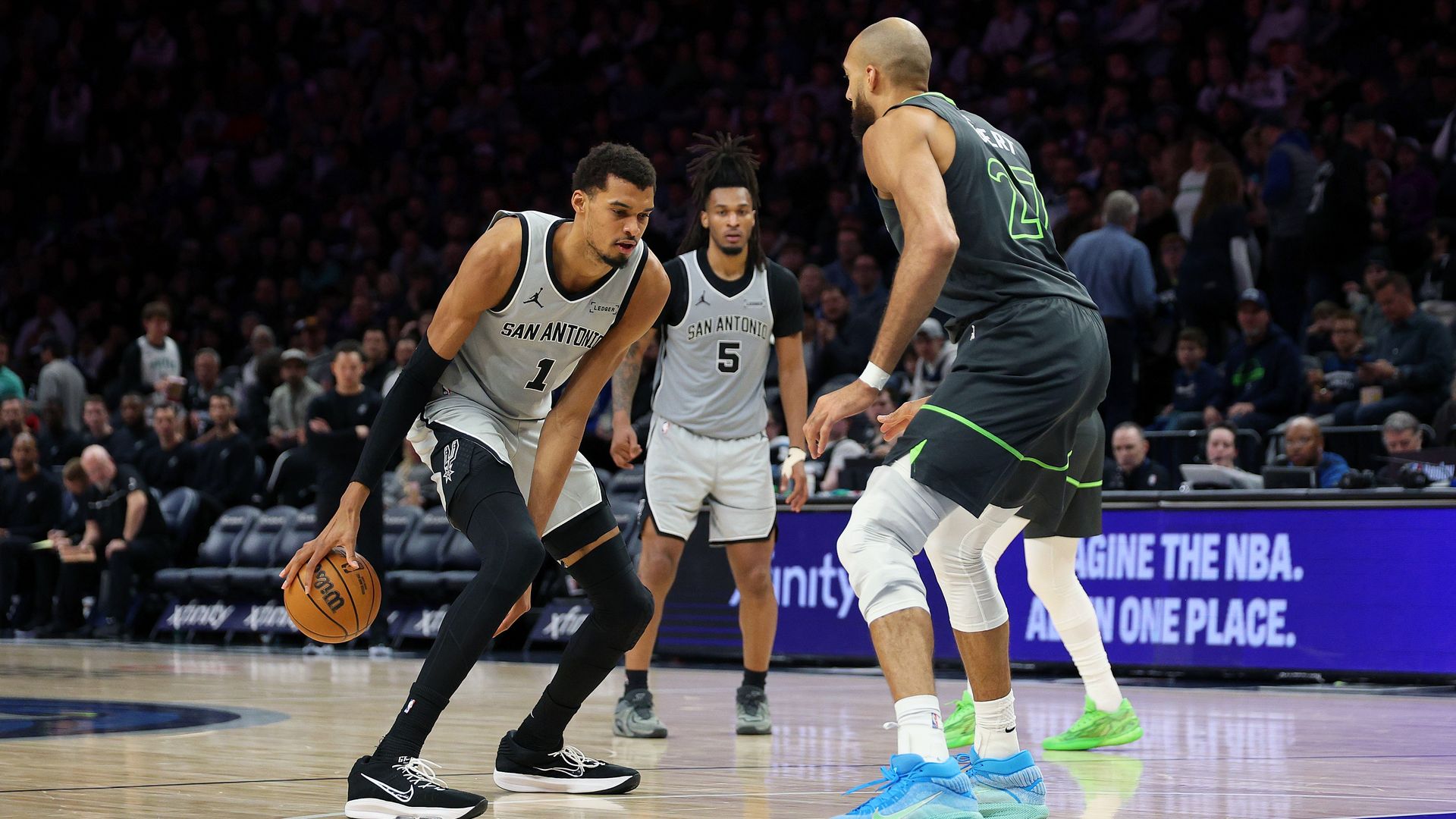 Spurs players in white jerseys drive the ball while a tall defender in gray with neon trim guards them; a packed arena watches from the stands.