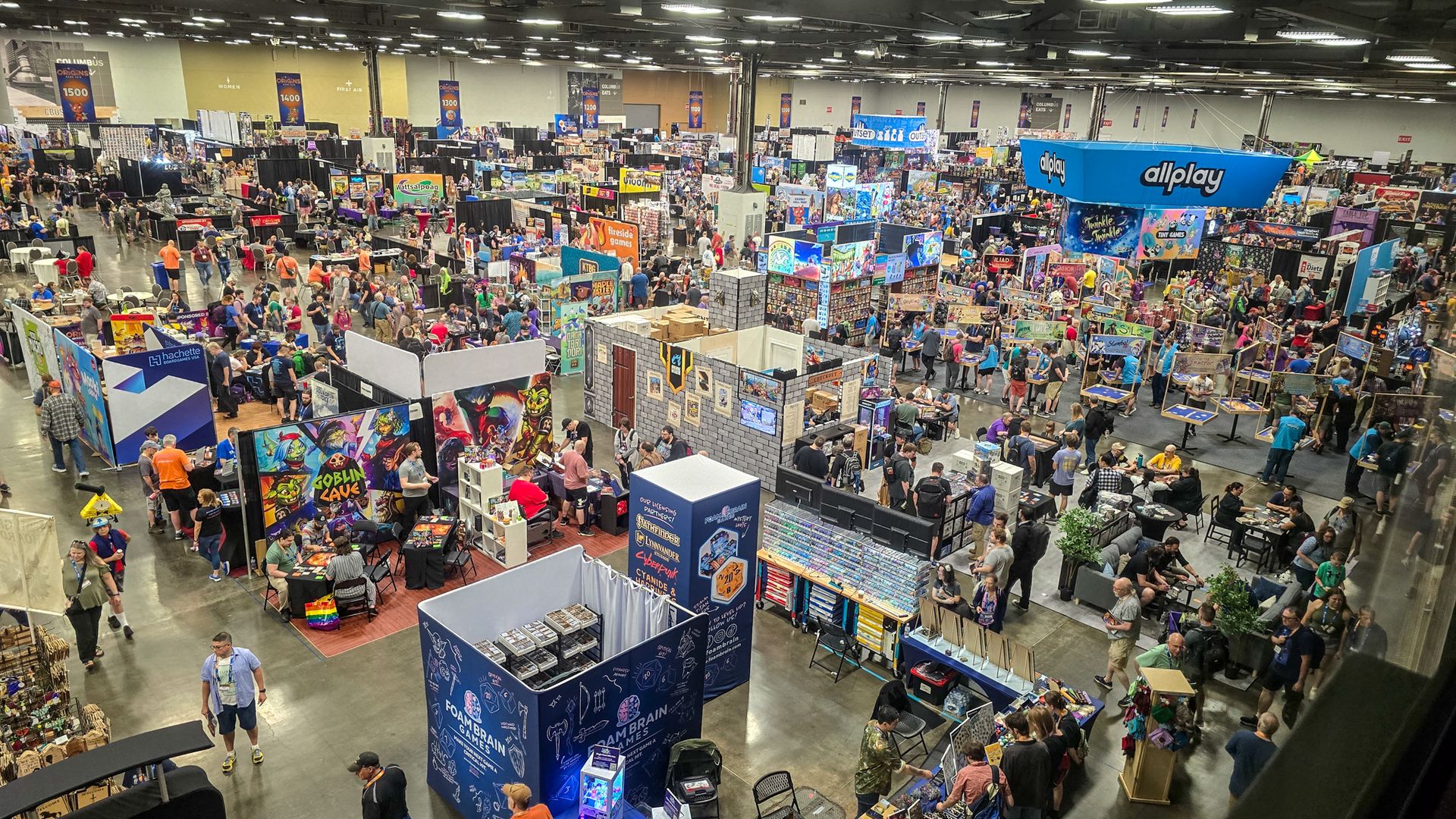 Panoramic view of a busy convention hall filled with colorful booths, posters, and a sea of attendees browsing games; a large blue allplay booth stands to the right.