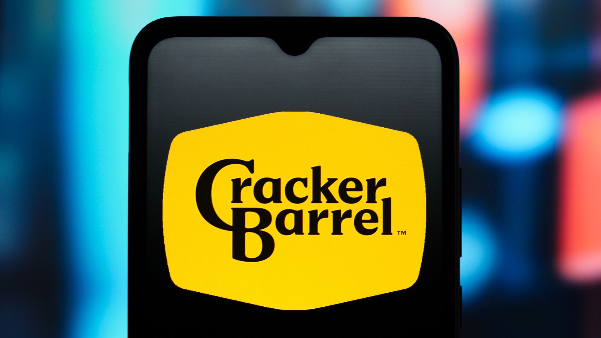 Yellow hexagonal logo with black text "Cracker Barrel" displayed on a smartphone against a blurred blue and red background.