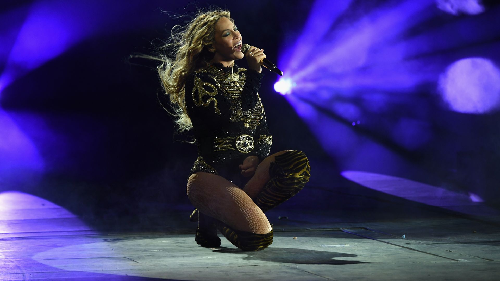 The performer Beyonce kneels on stage while singing into a microphone. She is wearing a black, sparkly bodysuit and is illuminated by purple lights.