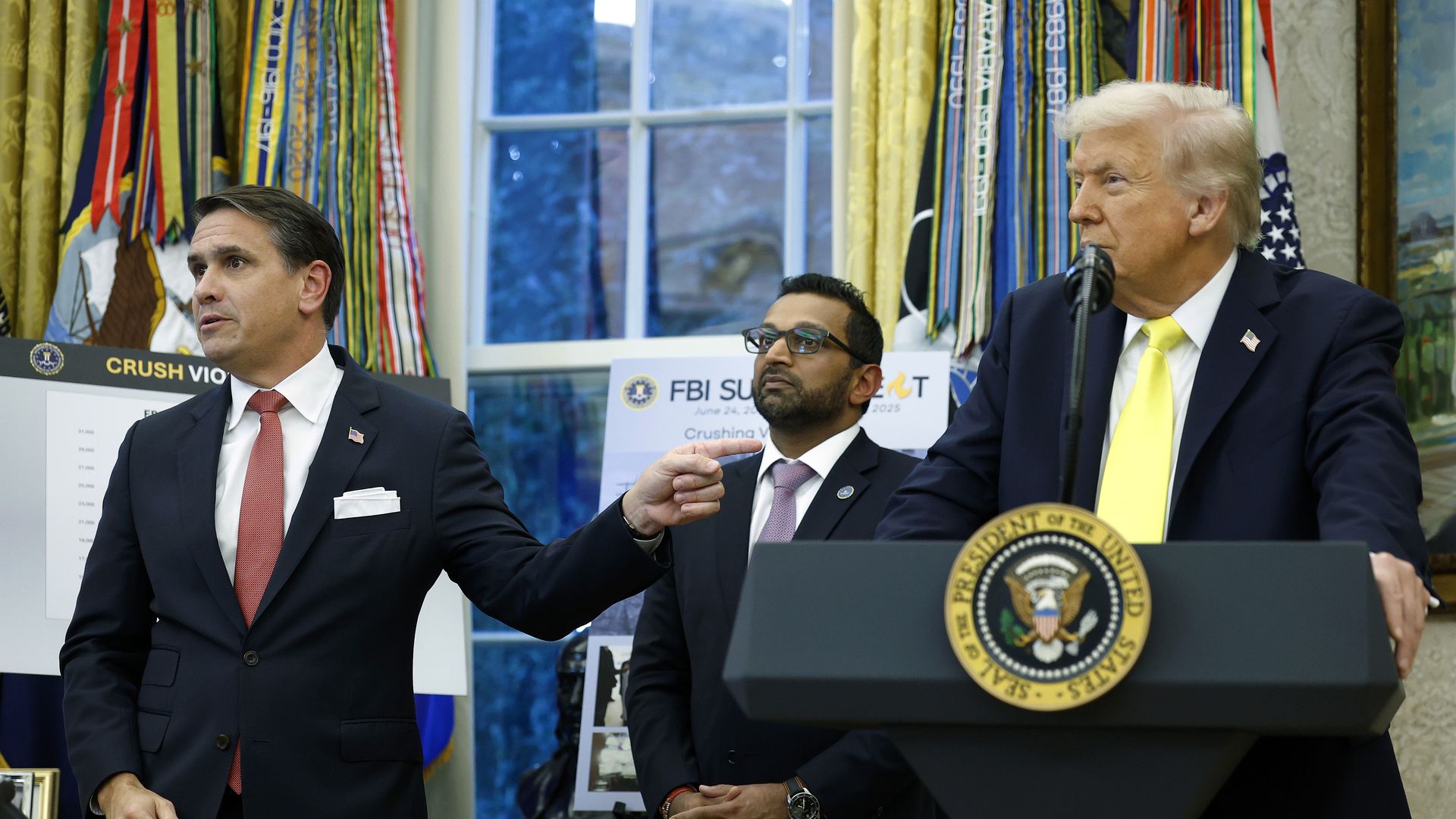 Todd Blanche — wearing a dark suit, a red tie and an American flag pin on his lapel — points to Donald Trump, who stands behind a podium while wearing a dark suit, yellow tie and an American flag pin on his lapel. Kash Patel can be seen standing between the two men.
