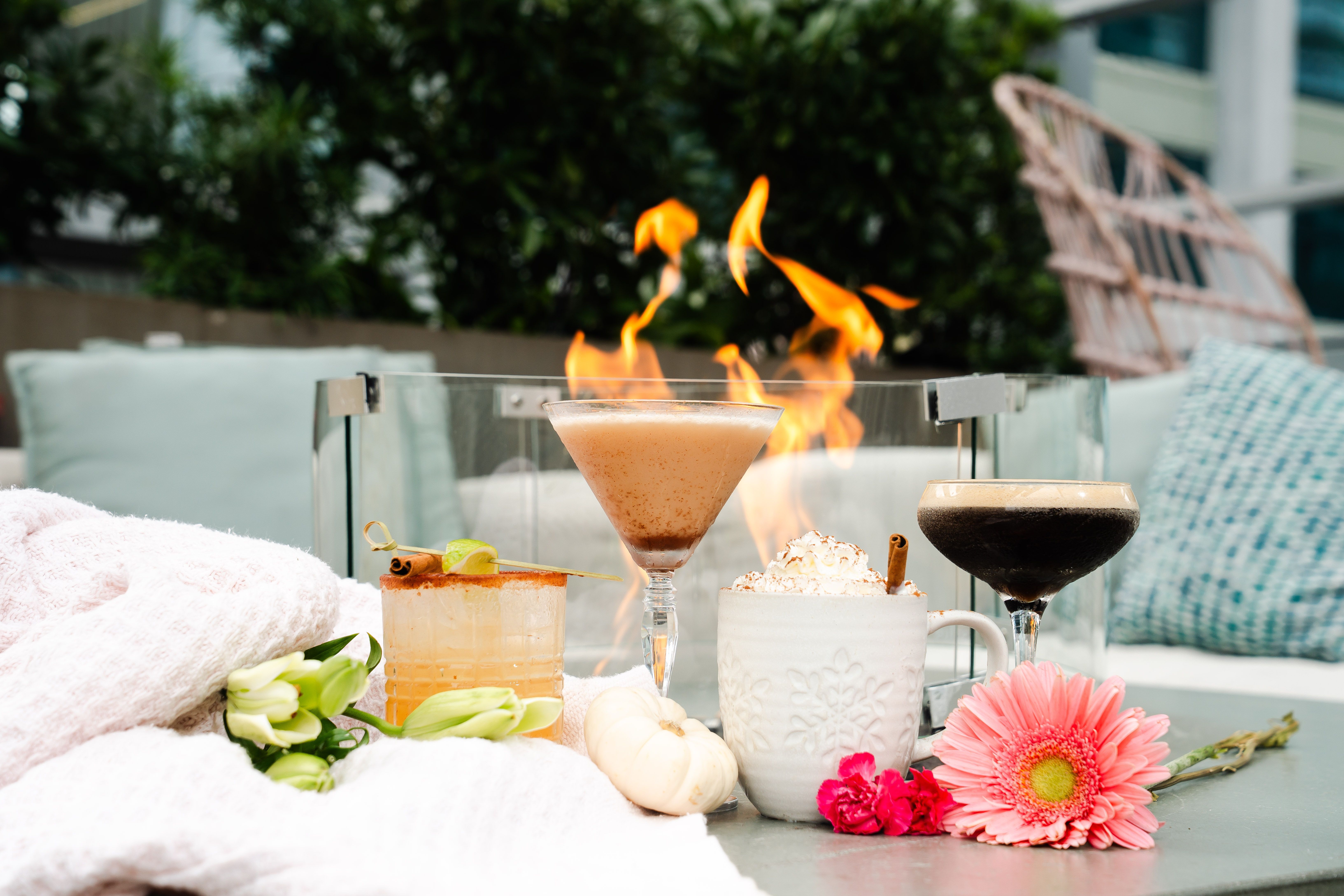 A cozy outdoor setting with four drinks on a table in front of a glass-enclosed fire pit, surrounded by white knit blankets, flowers, and a small white pumpkin.