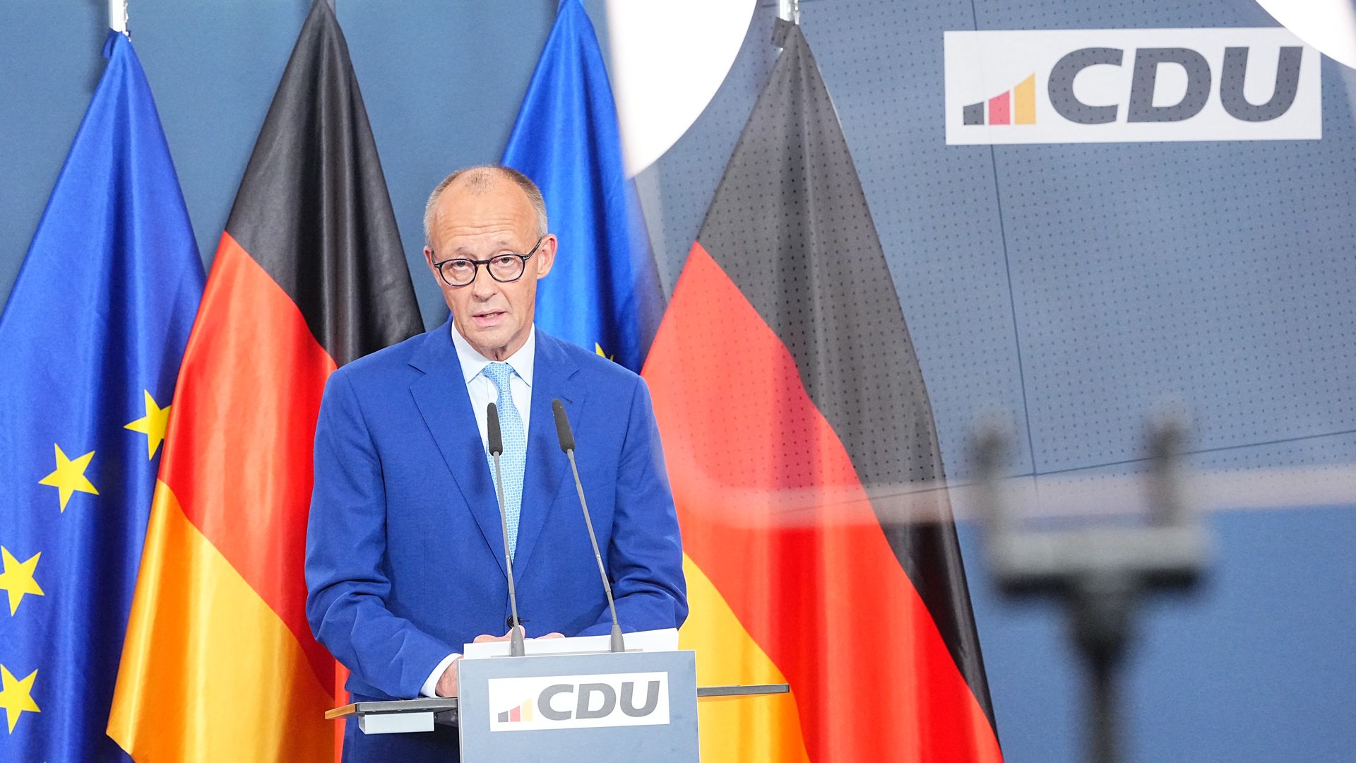 A man in a blue suit and glasses speaks at a podium with two microphones, flanked by European and German flags and a CDU banner in the background.