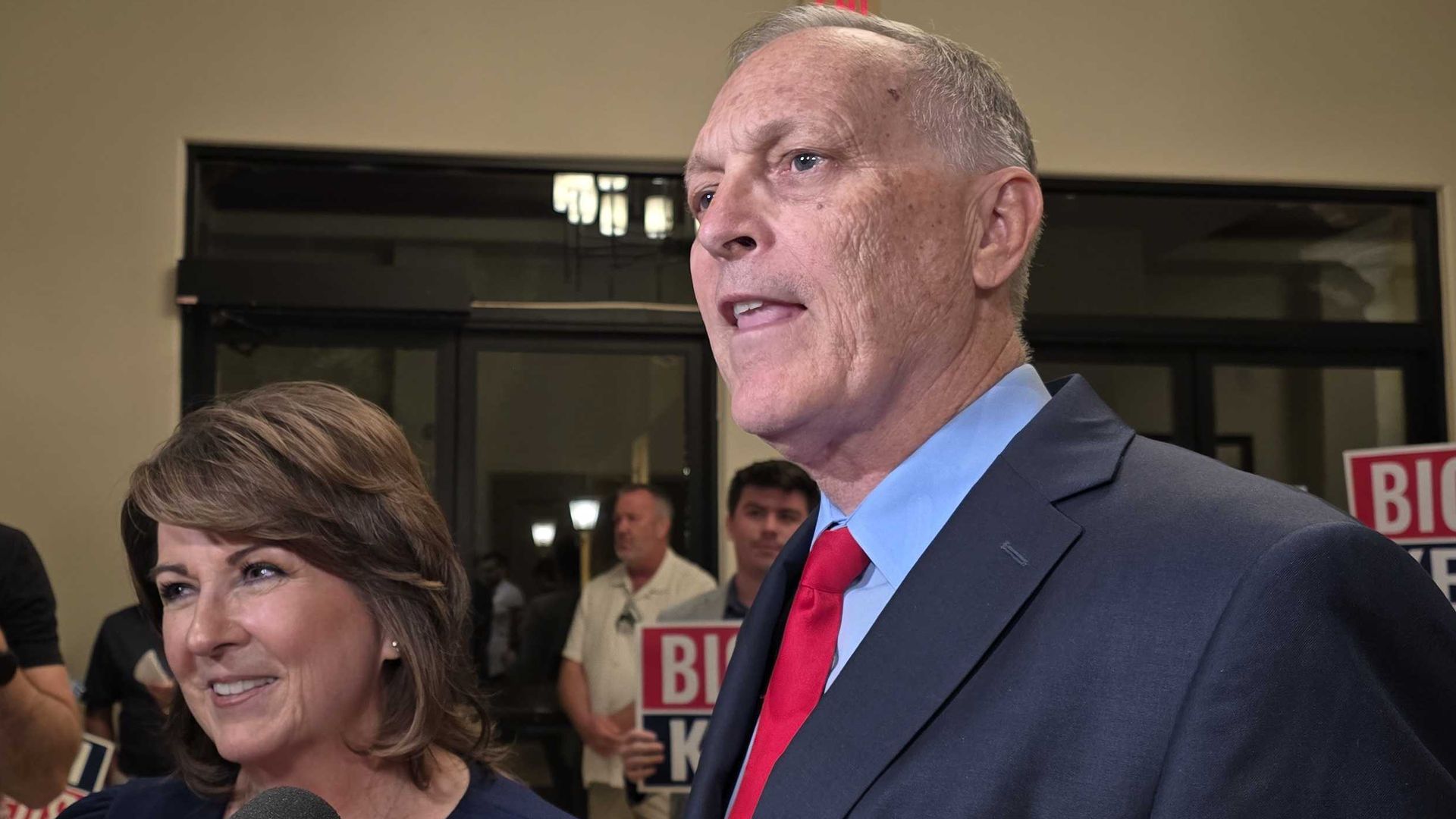 Andy Biggs in a navy suit with a red tie speaks to reporters at an indoor campaign event, beside a smiling Sine Kerr; microphones fill the foreground and BIGGS signs are visible in the background.