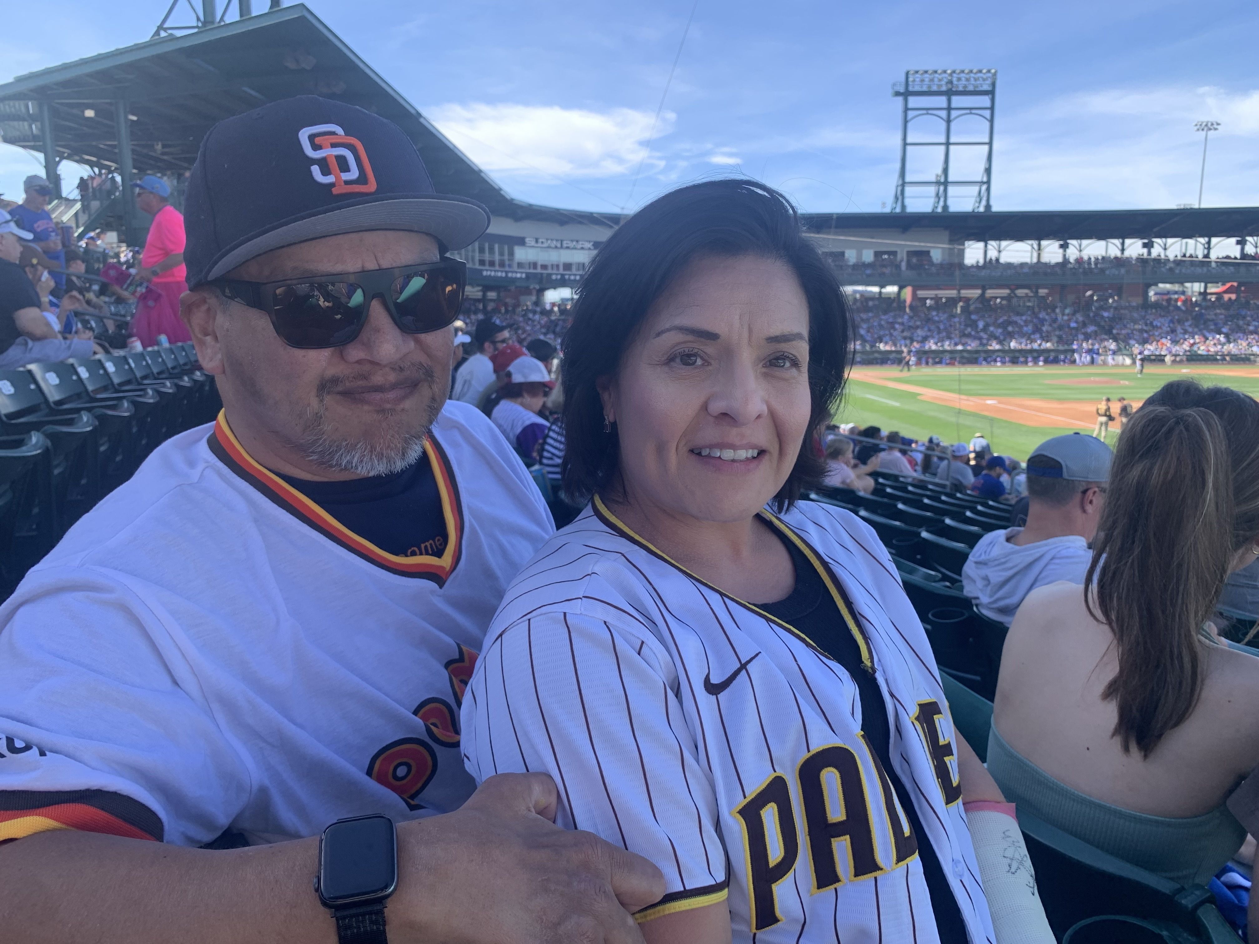 A man and woman sit together wearing San Diego Padres white baseball jerseys at a sunny stadium with a baseball game in progress behind them.