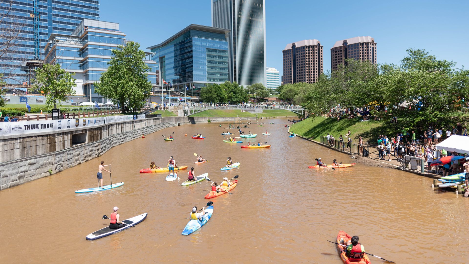 Wide city riverfront with muddy brown water, numerous kayakers and paddleboarders, a grassy embankment crowd, and modern glass skyscrapers beneath a clear blue sky; a construction crane visible on the left.
