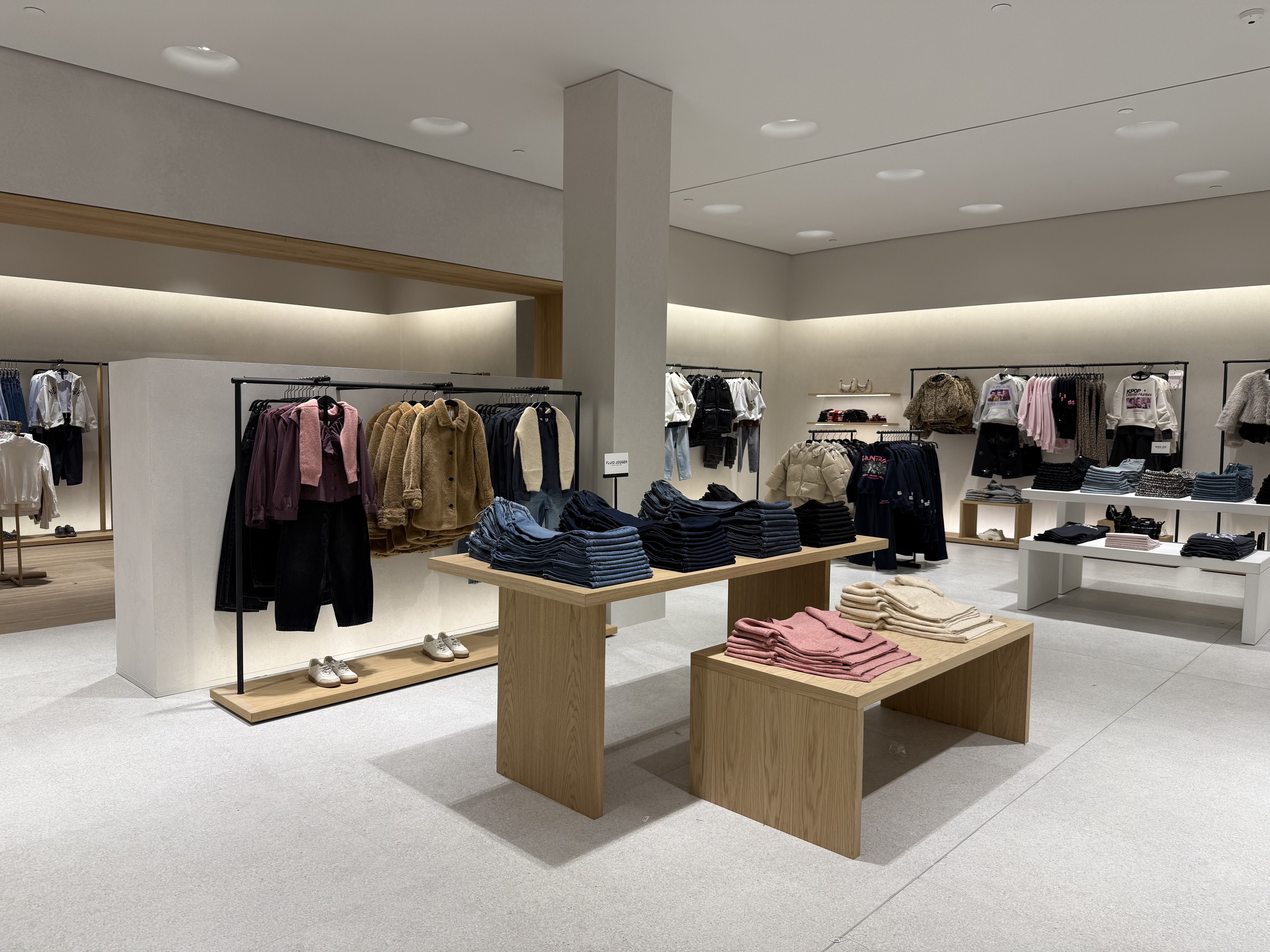 Modern clothing store interior with beige walls, light grey floor, and wooden display tables. Clothes include folded sweaters in pink and beige, denim jeans, and racks of jackets and sweaters.