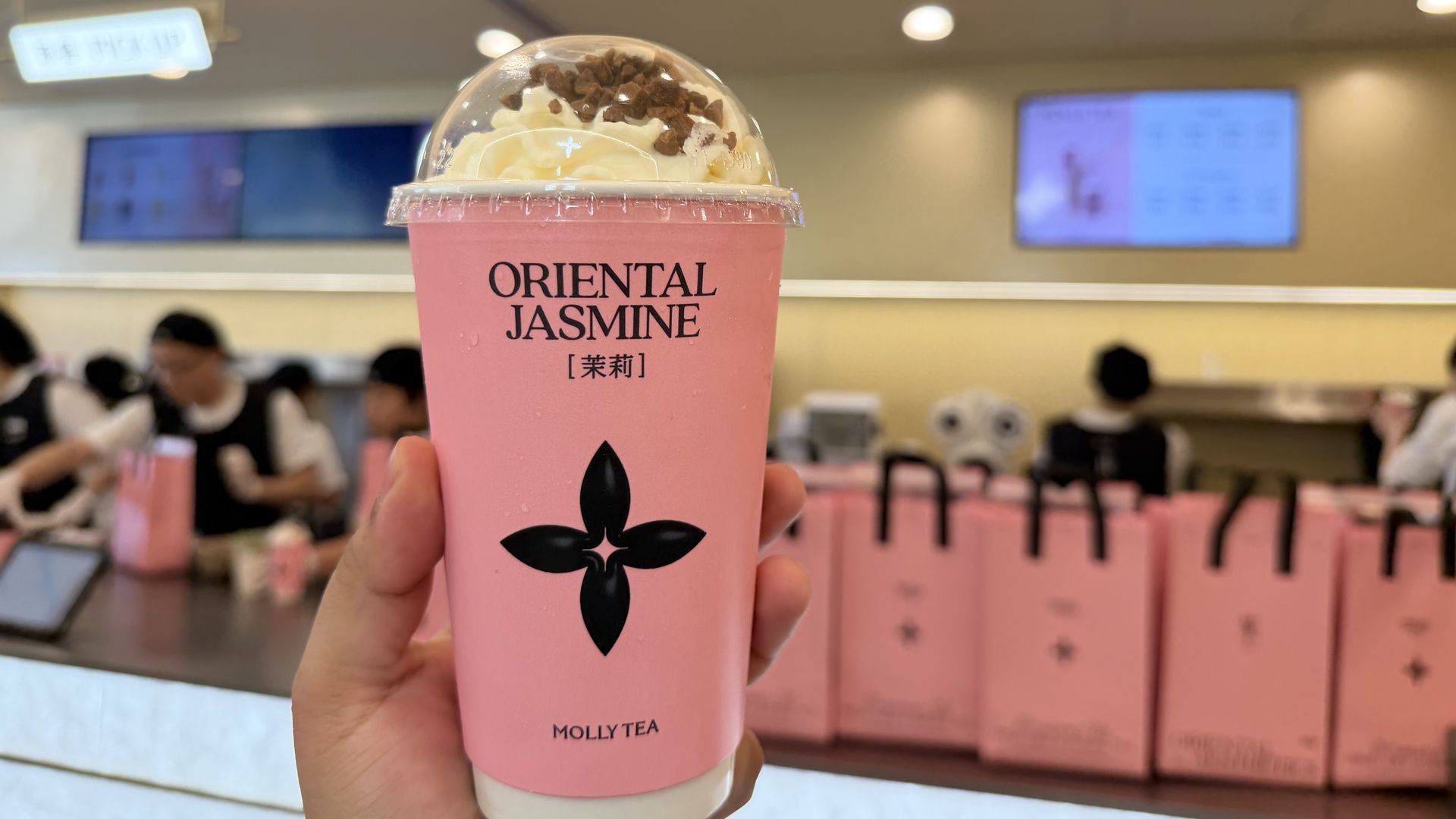 Hand holding a pink cup of Oriental Jasmine tea with whipped cream and chocolate bits on top inside a tea shop with pink bags and people in the background.