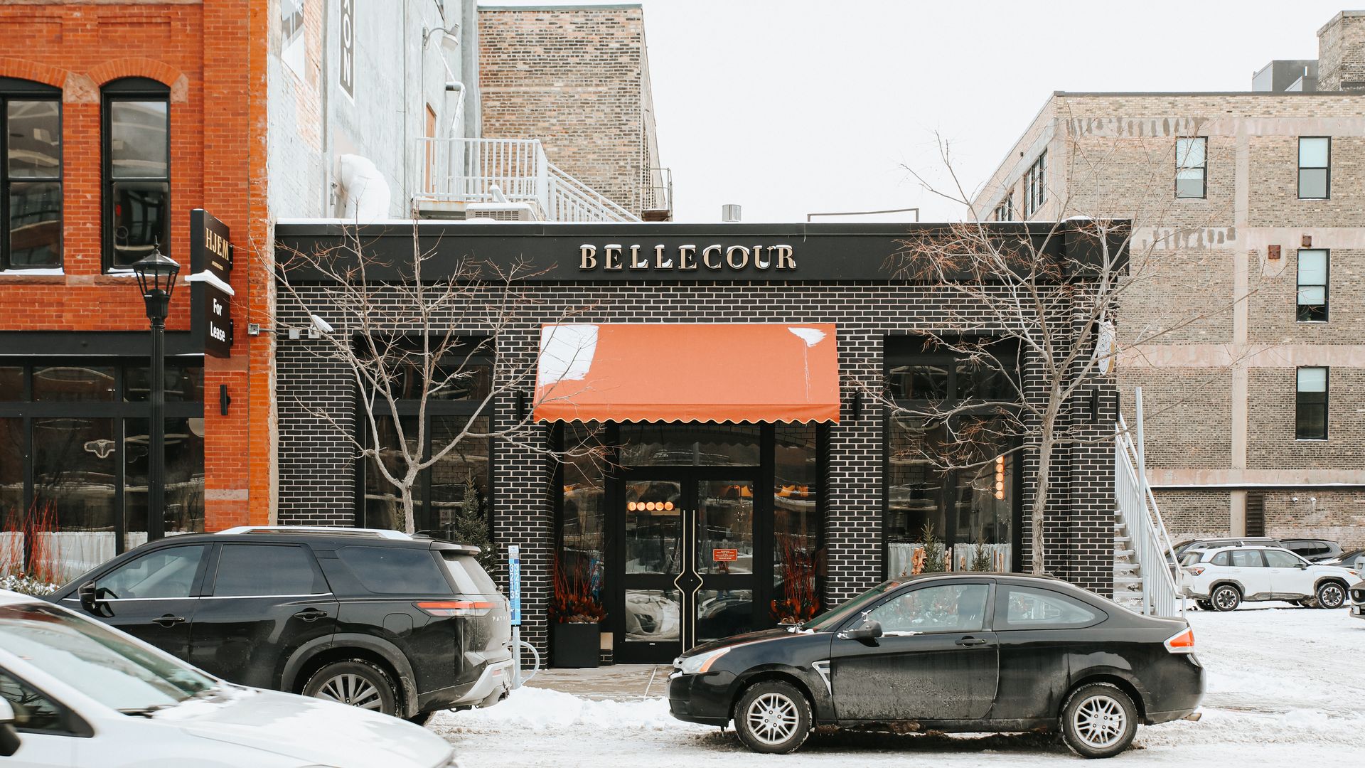 Black brick building with orange awning and BELLECOUR sign above entrance, snowy street with parked black and white cars, leafless trees, and adjacent red and beige brick buildings.