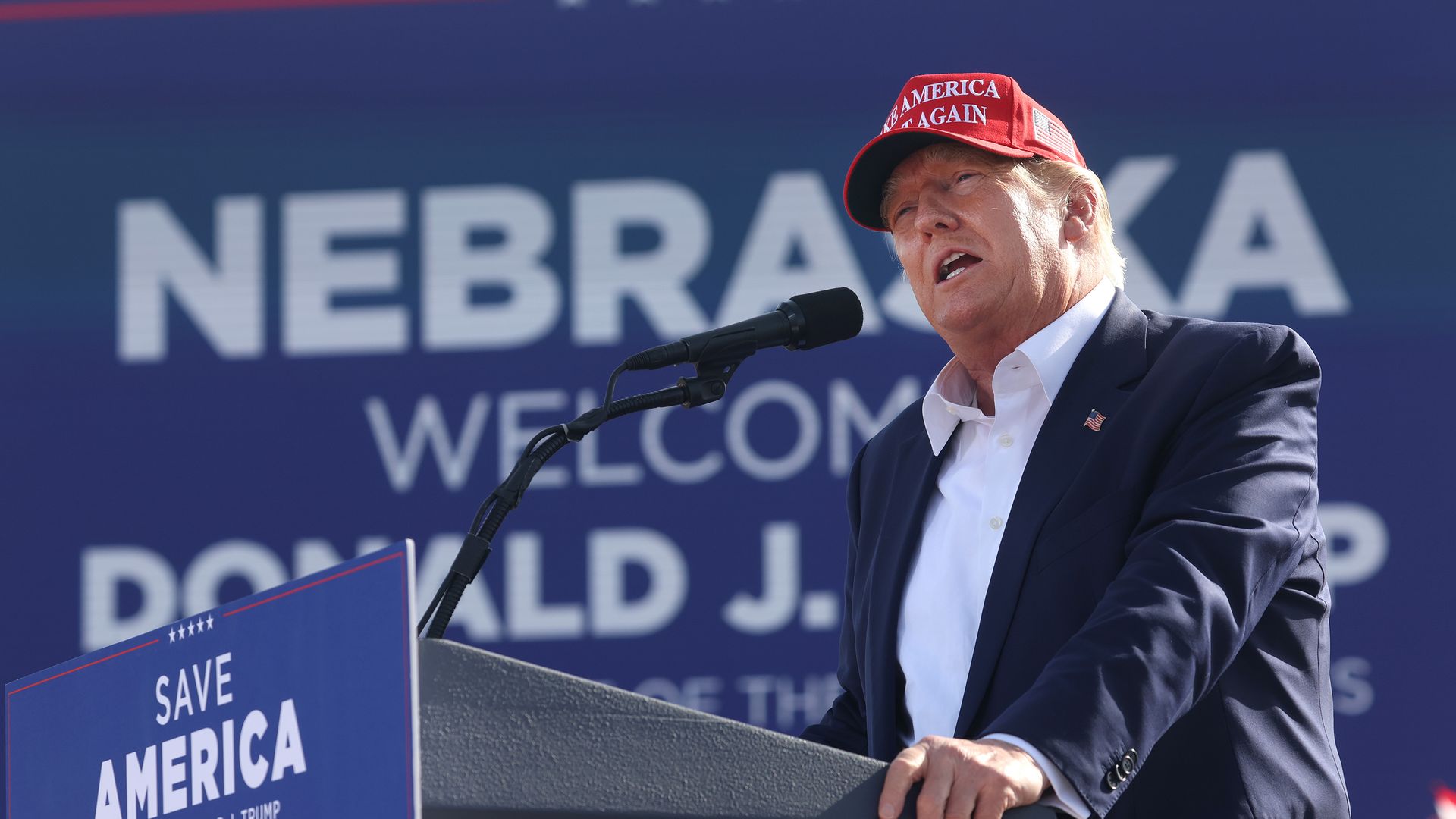 Photo of Donald Trump speaking from a podium and wearing a red MAGA hat