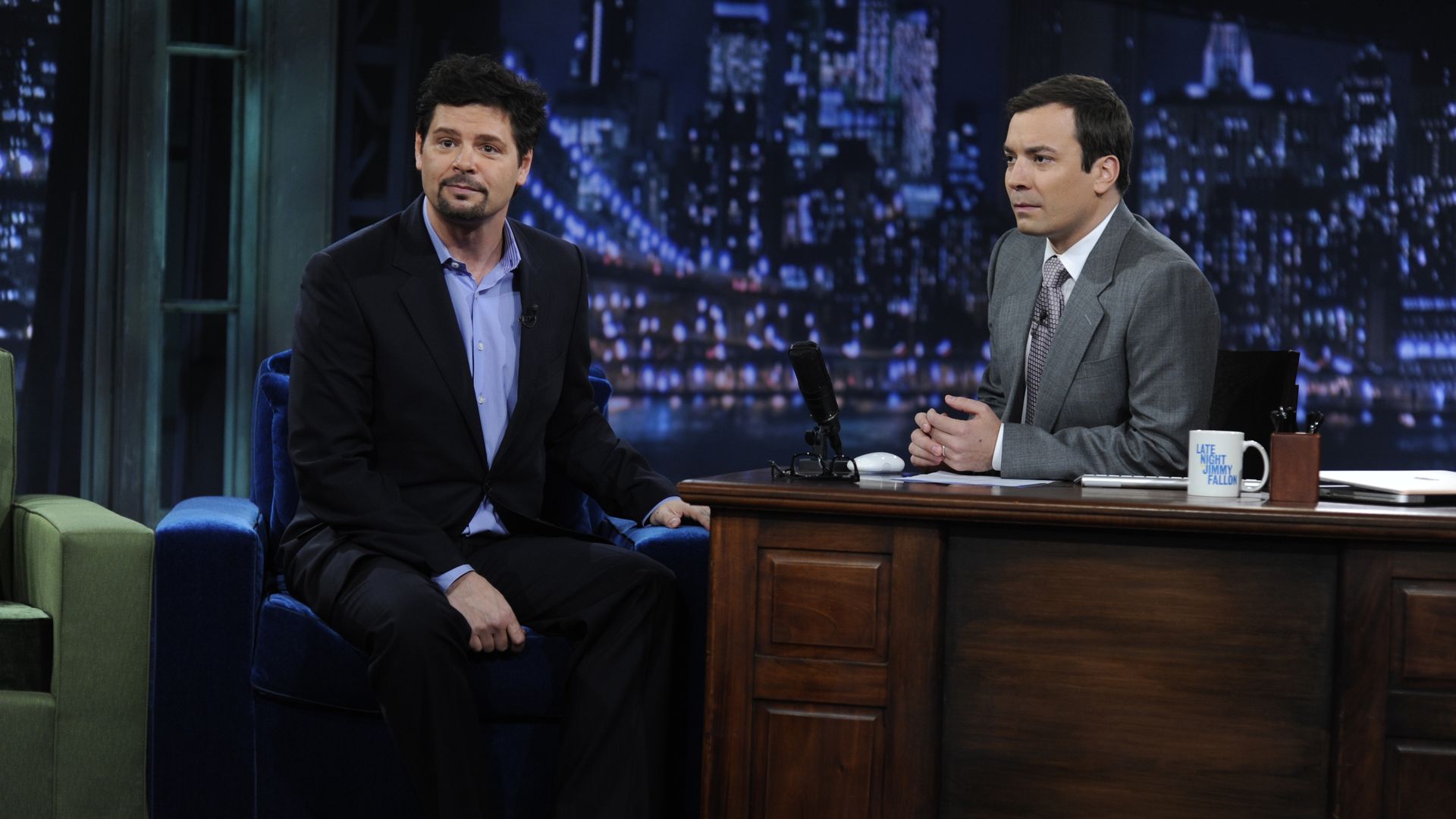 Photo of two men talking on a television set with a desk and a couch 