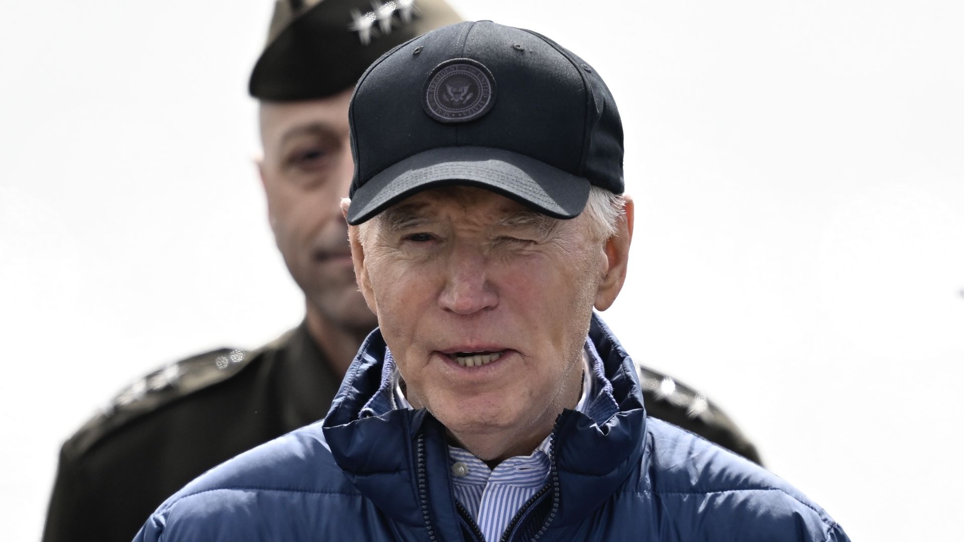 President Biden, wearing a blue puffer jacket, white and blue striped shirt and black hat with the presidential seal, stands in front of a soldier outside.