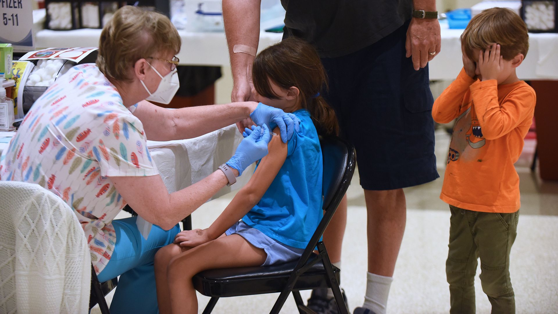 Picture of a nurse putting a vaccine on a little girl while her brother covers his eyes