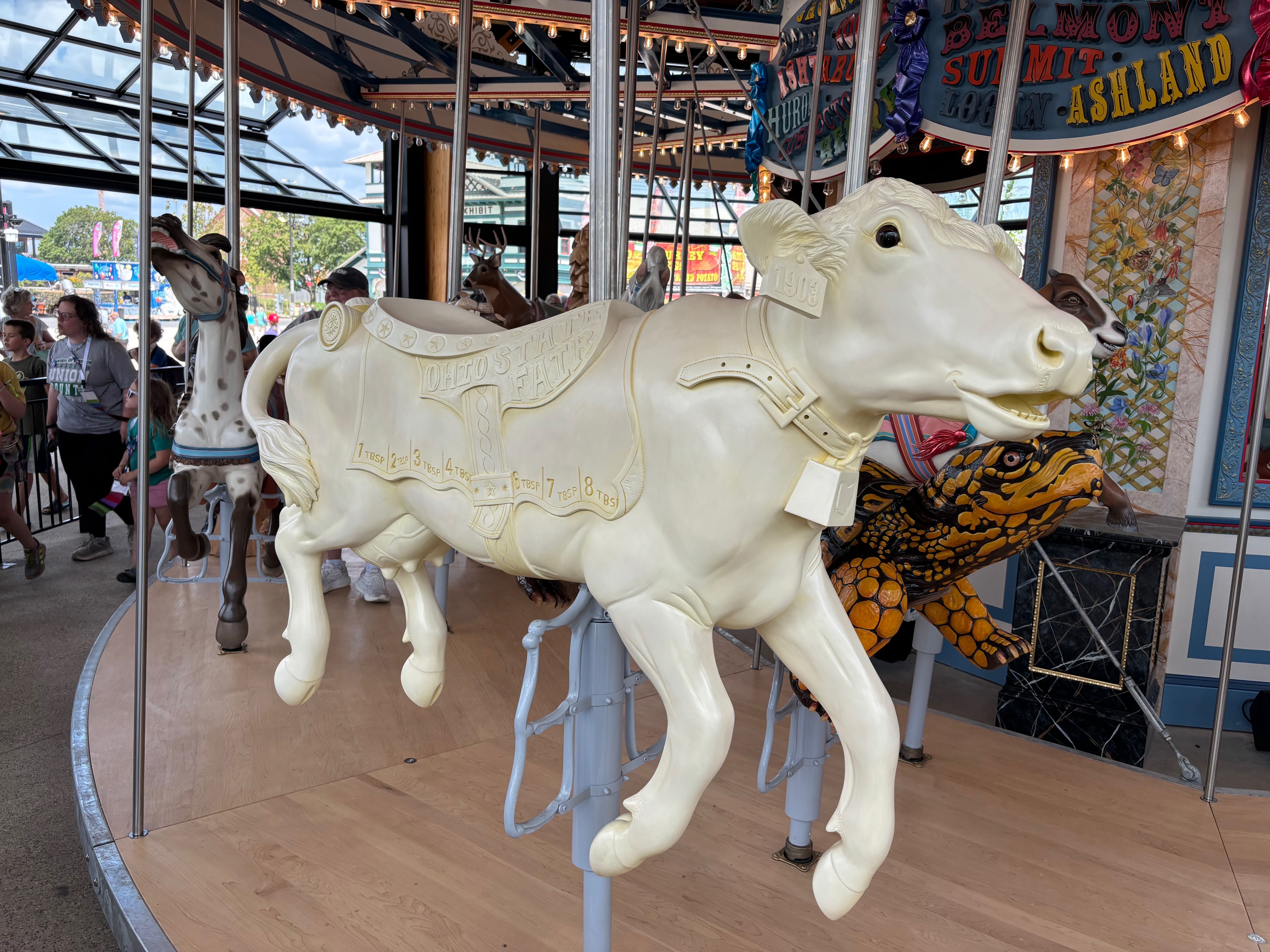 Carousel animals: a large white cow in front with detailed saddle, harness, and carved features; other animals and riders visible behind, under string lights in a glass-roofed pavilion.
