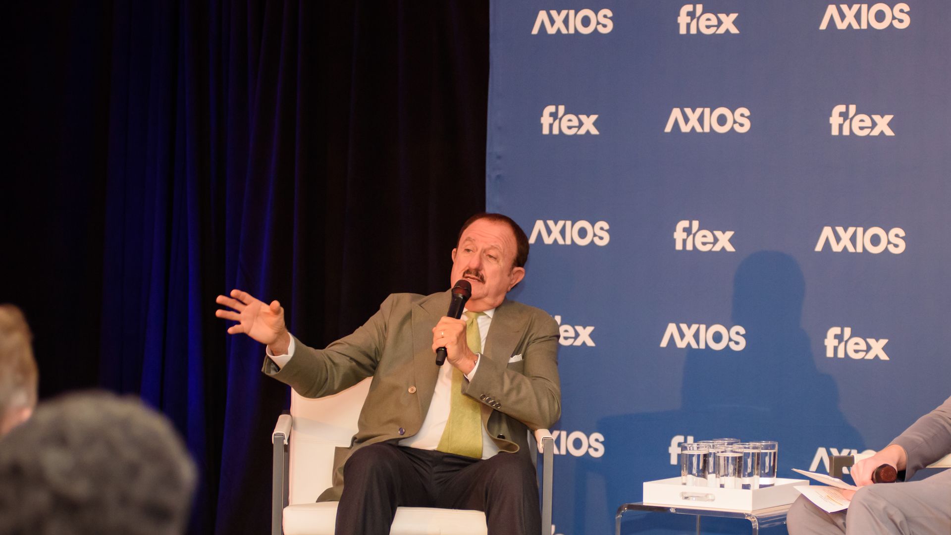 Man in green tie and brown suit speaking into a microphone on stage at an Axios Flex event, gesturing with his left hand, seated on a white chair with blue backdrop.
