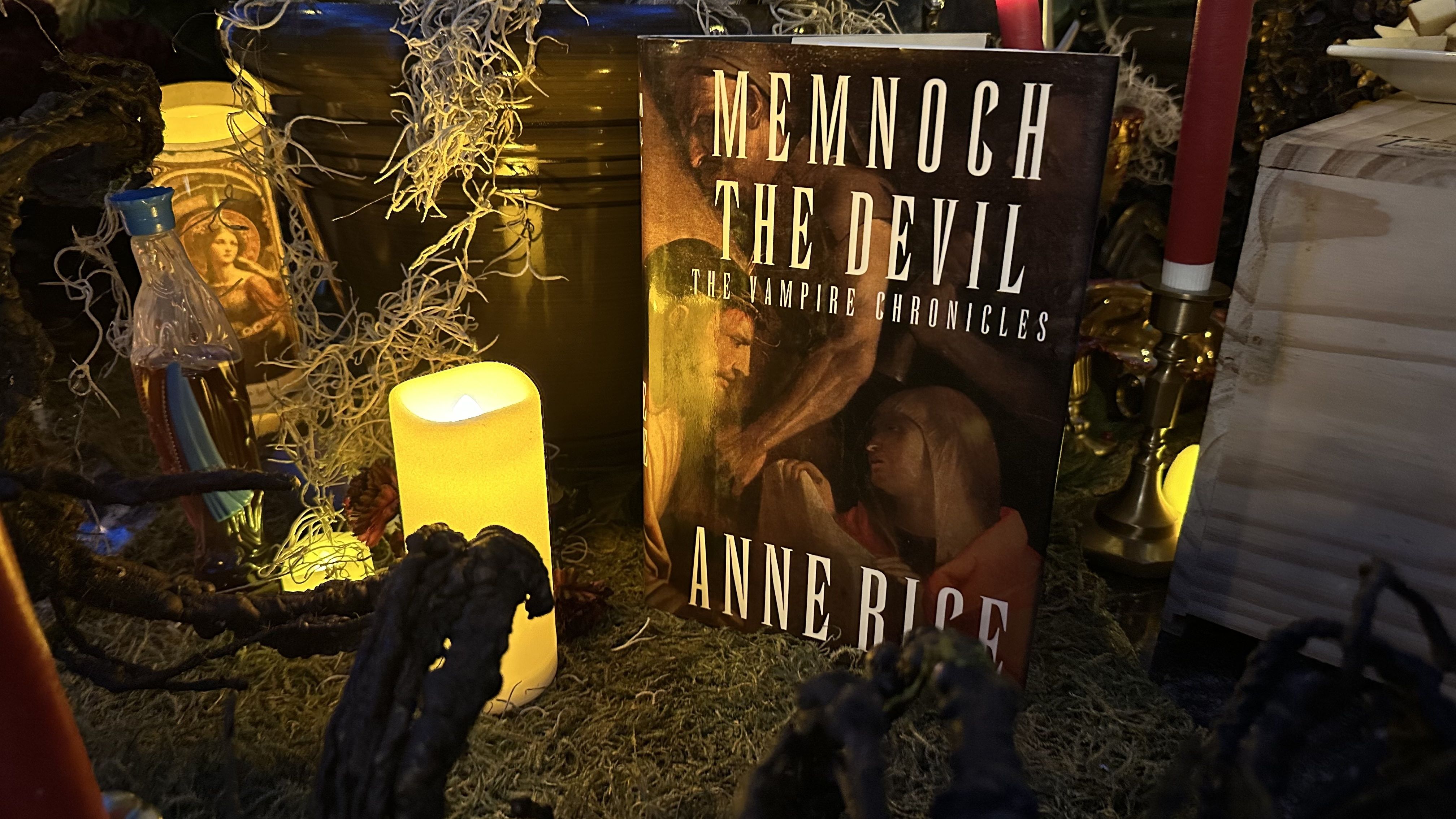Book titled "Memnoch The Devil" by Anne Rice from The Vampire Chronicles, surrounded by a glowing yellow candle, dried moss, a red candle on a brass holder, and decorative dark branches.