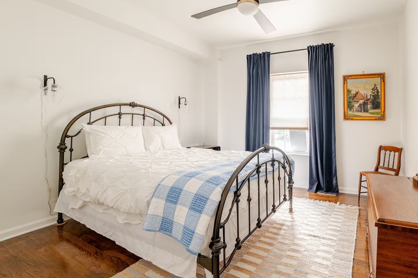 Bright bedroom with white walls, a black metal bed frame, white bedding, a blue and white checkered throw, dark blue curtains, hardwood floor, wooden furniture, and a framed landscape painting.