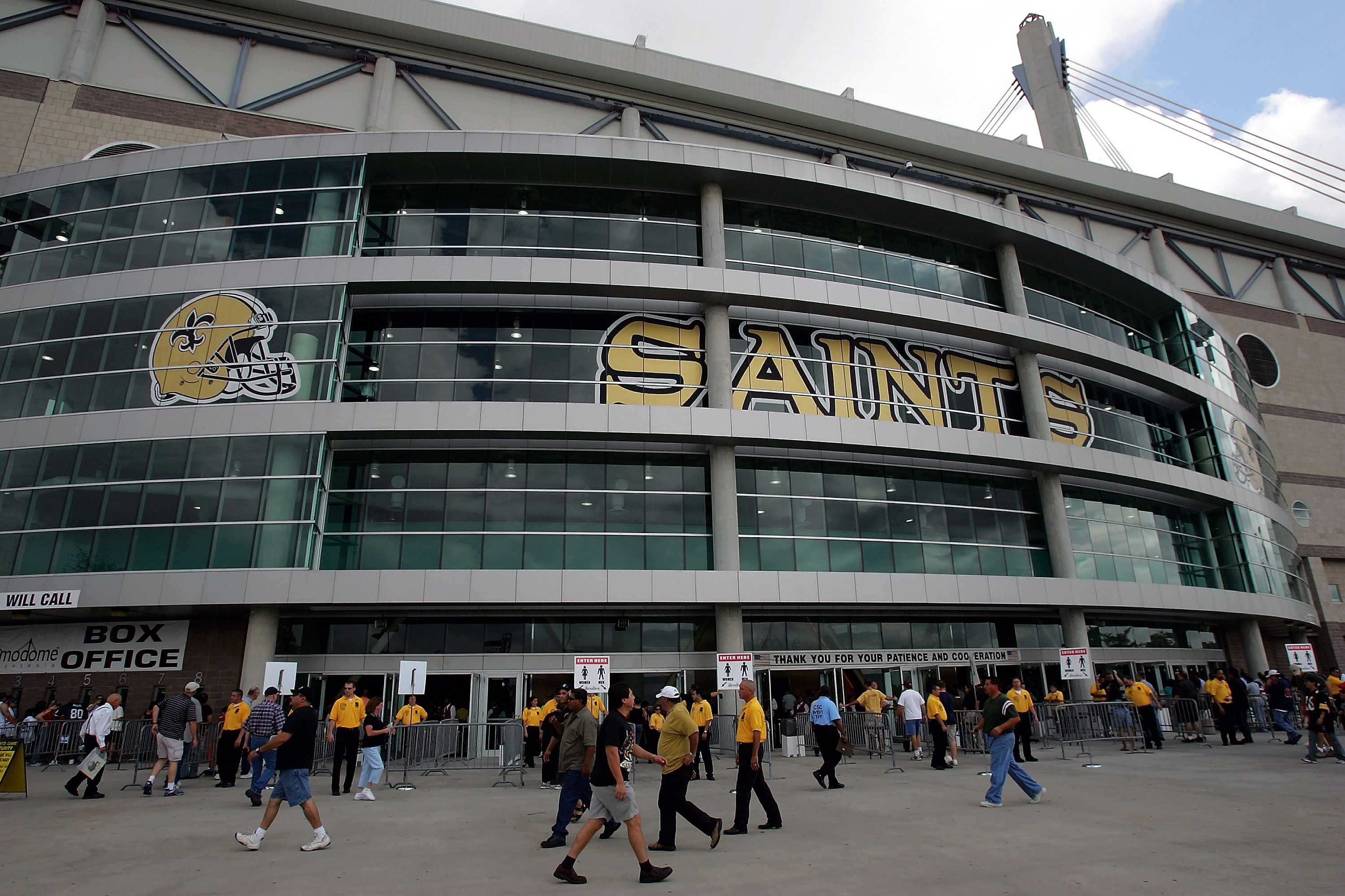 Exterior of a large stadium with glass windows displaying a gold football helmet and the word "SAINTS" in gold. People walk and security personnel in yellow shirts stand near entrances.