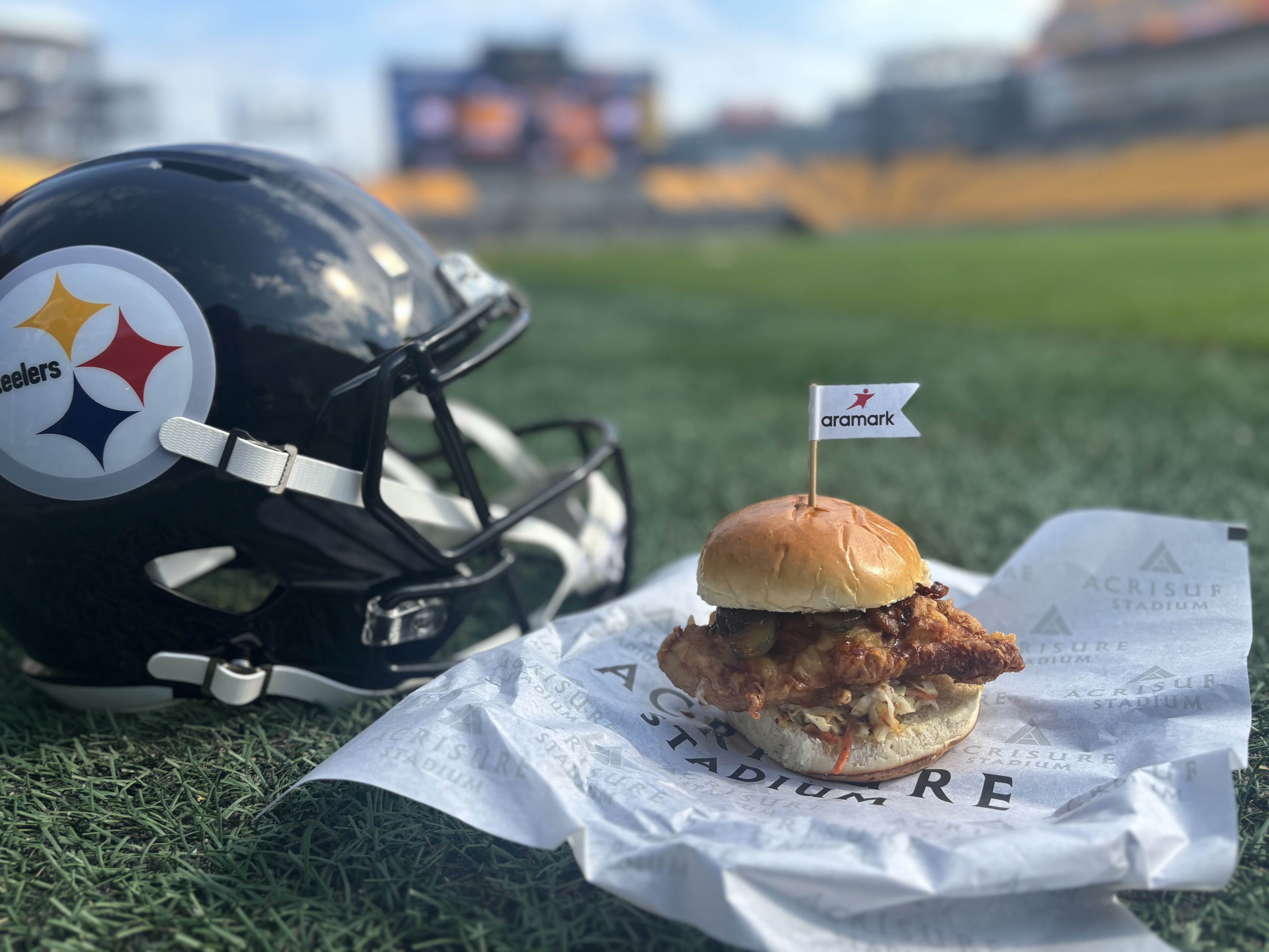 A Pittsburgh Steelers black football helmet on artificial turf beside a crispy chicken sandwich with mustard topped with an Aramark flag, at Acrisure Stadium.