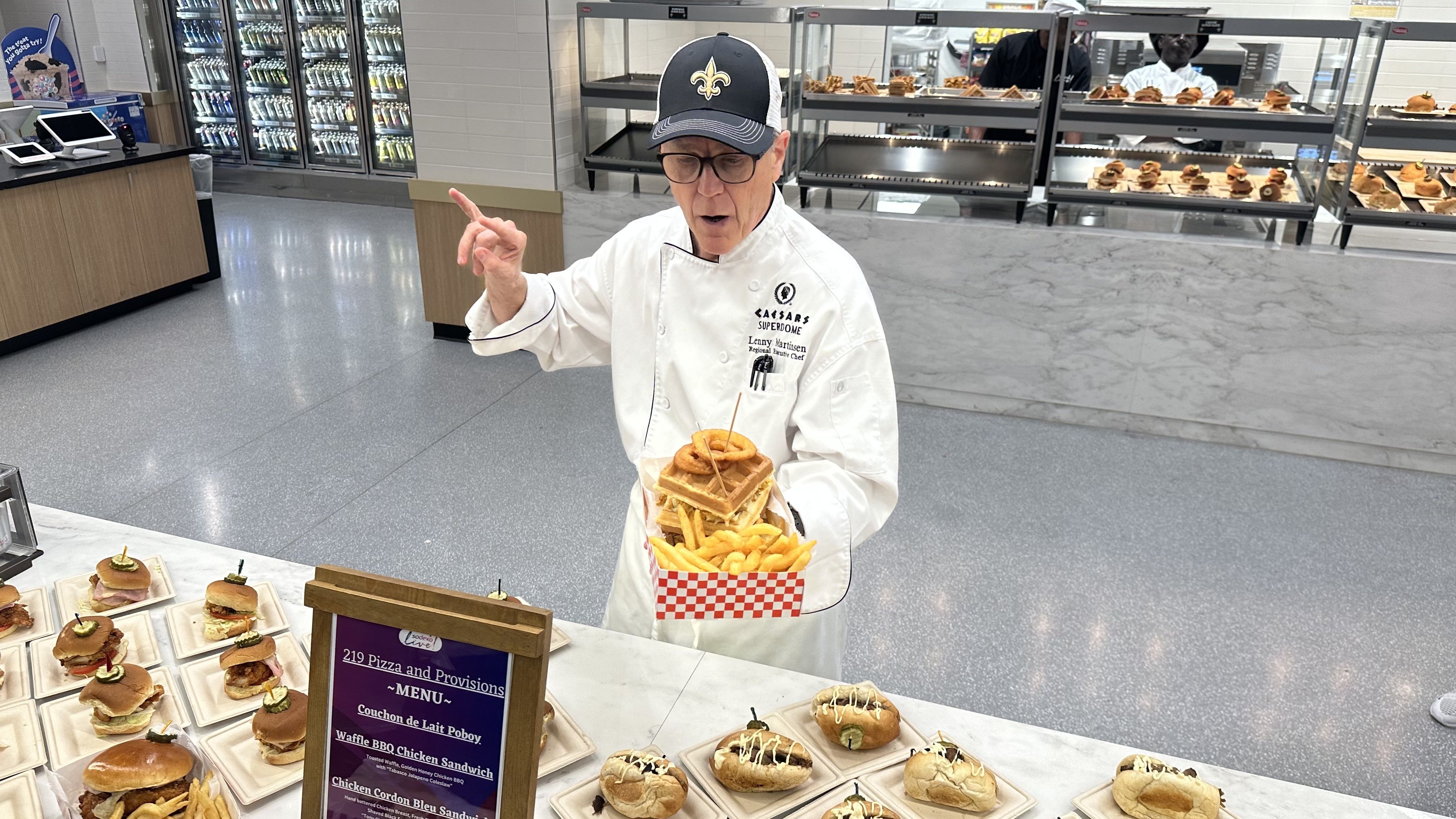 A man holds up a waffle sandwich and fries. He is wearing a chef's coat and a Saints hat.