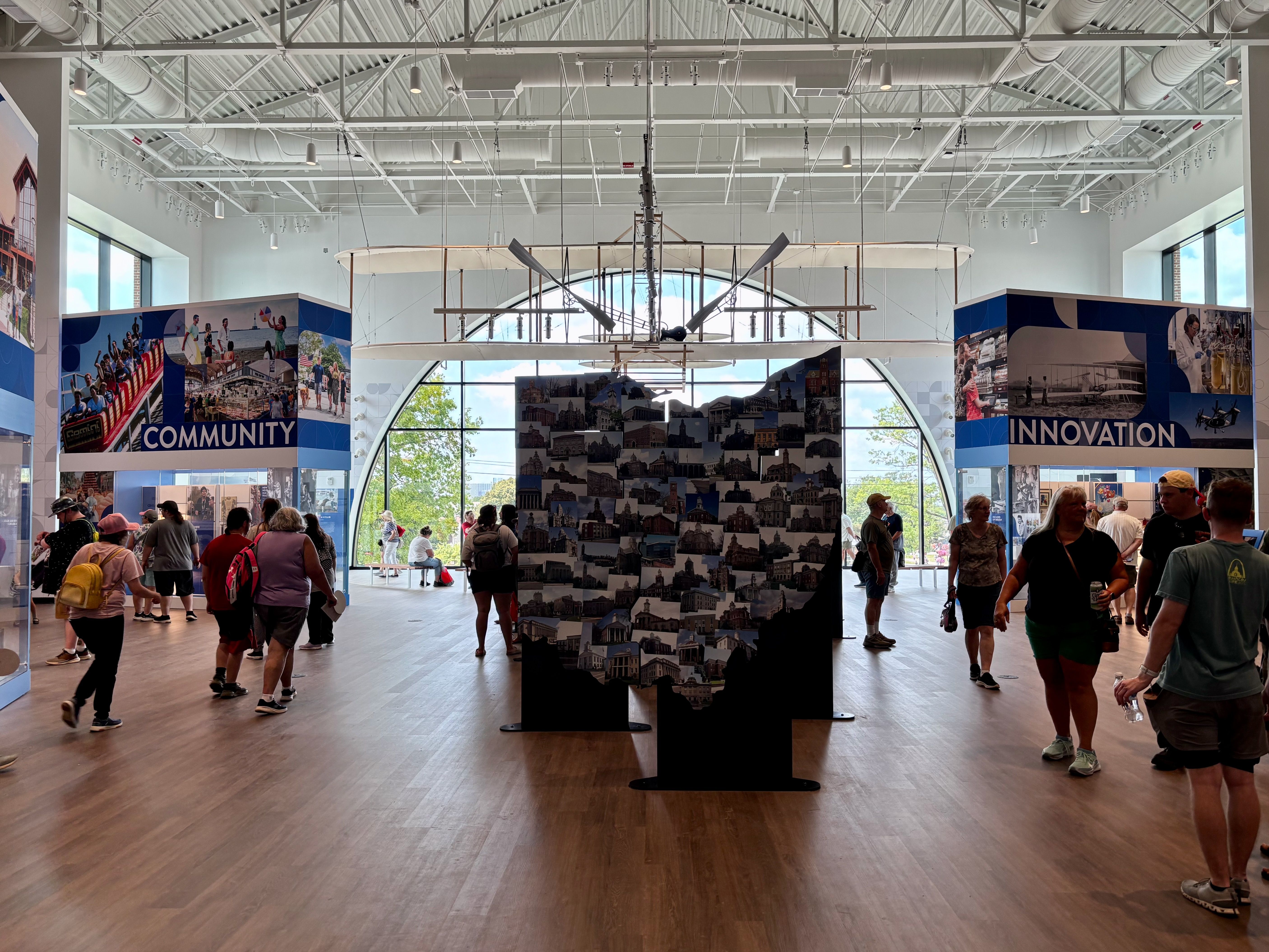 Inside a bright museum hall, a suspended vintage airplane hangs from a high white ceiling. Visitors walk past blue panels reading "COMMUNITY" and "INNOVATION" with a large arched window behind.