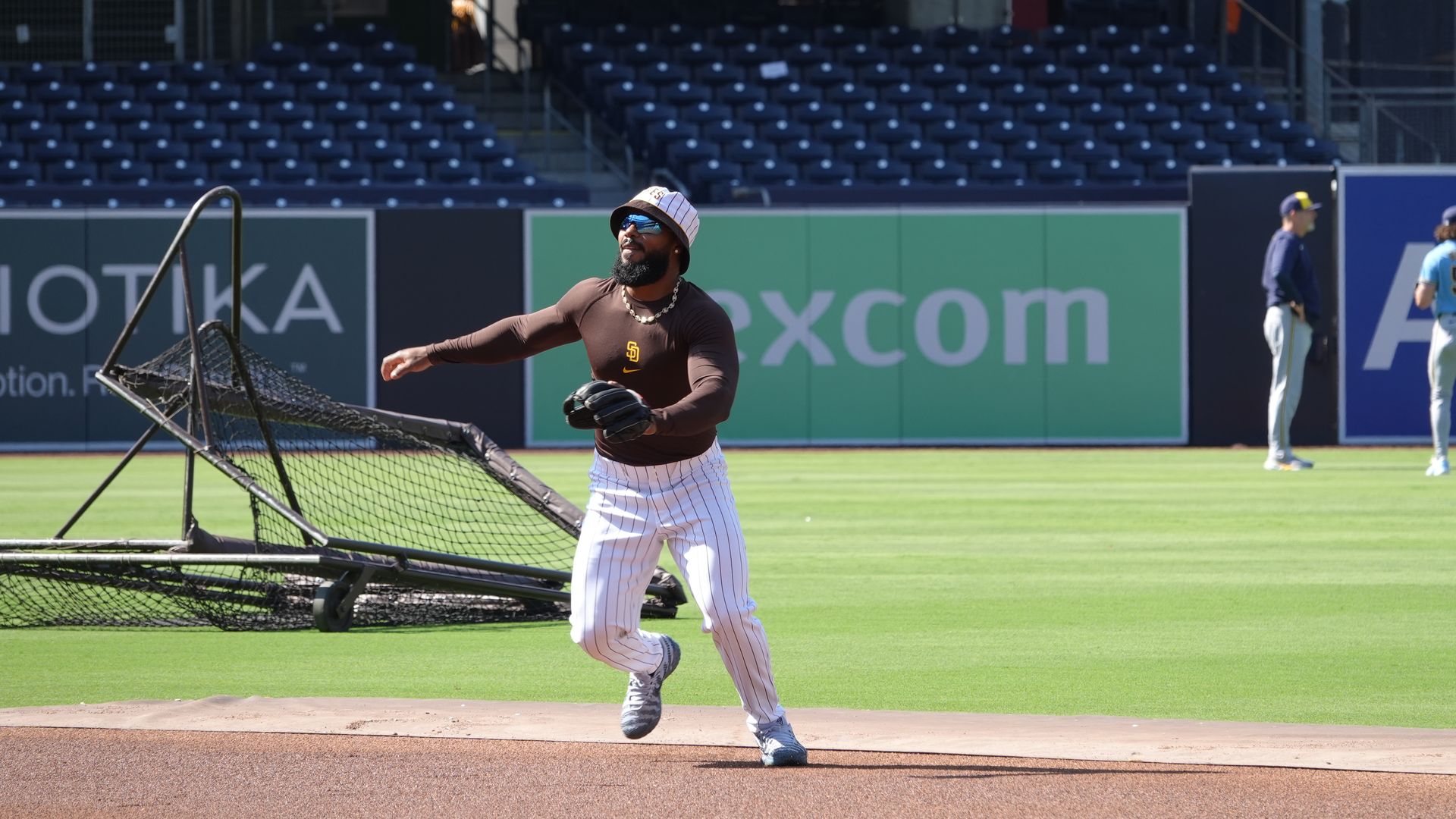Baseball pitcher in a brown shirt, white pinstripe pants, striped bucket hat, sunglasses and a necklace, throwing near a protective net on a sunny field; empty blue stadium seats behind.