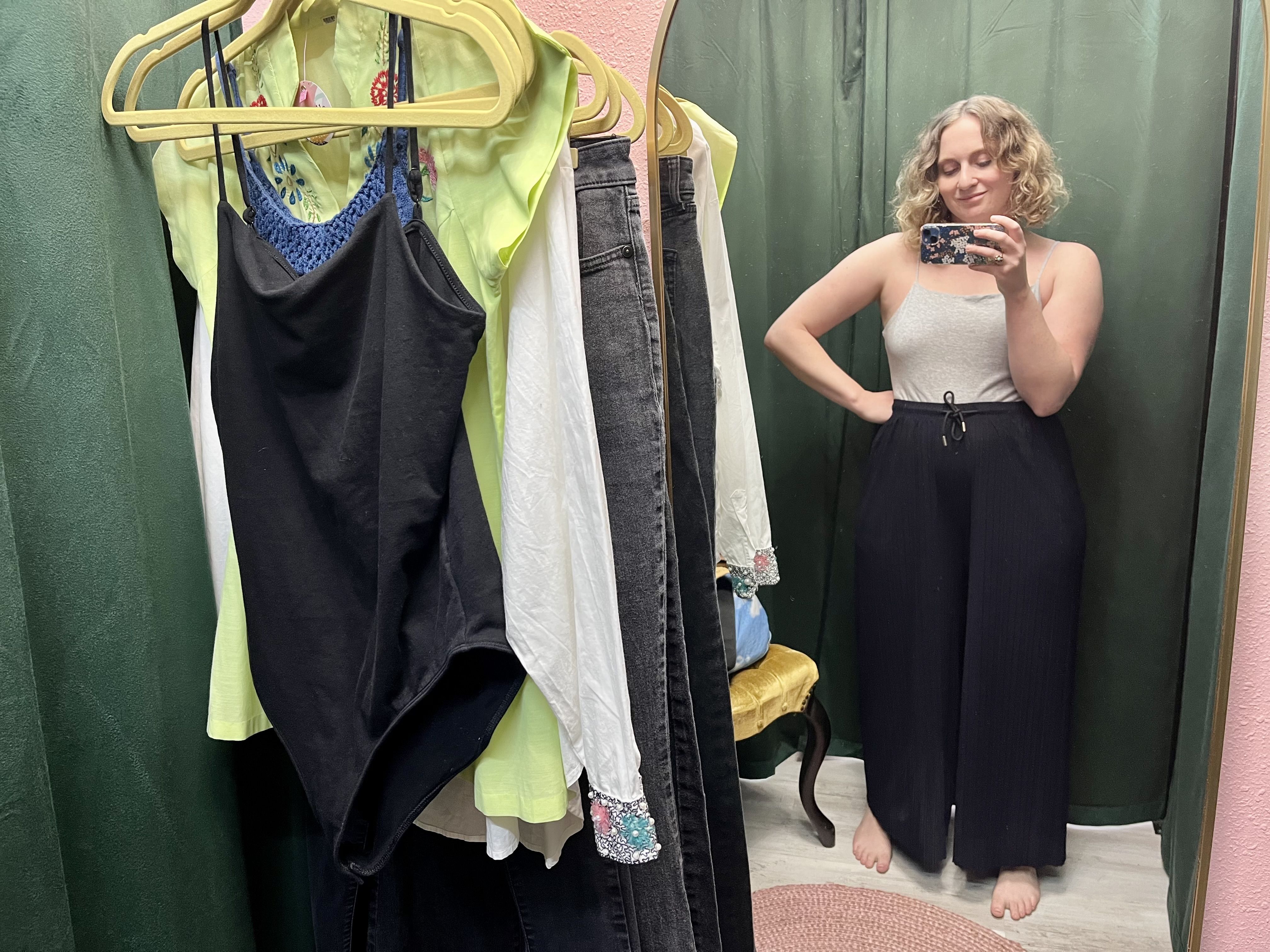 A woman in wide-leg navy pants and a gray top taking a mirror selfie in a dressing room next to a rack of clothes.