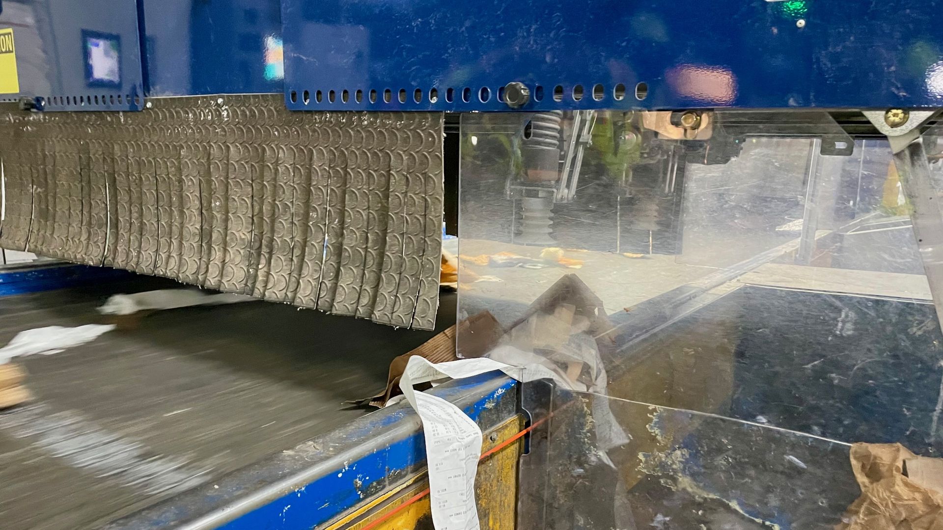 Blue industrial machine with a perforated metal base and a textured conveyor belt. A long receipt-like paper hangs over the edge, while clear guards and metal panels enclose the workspace.