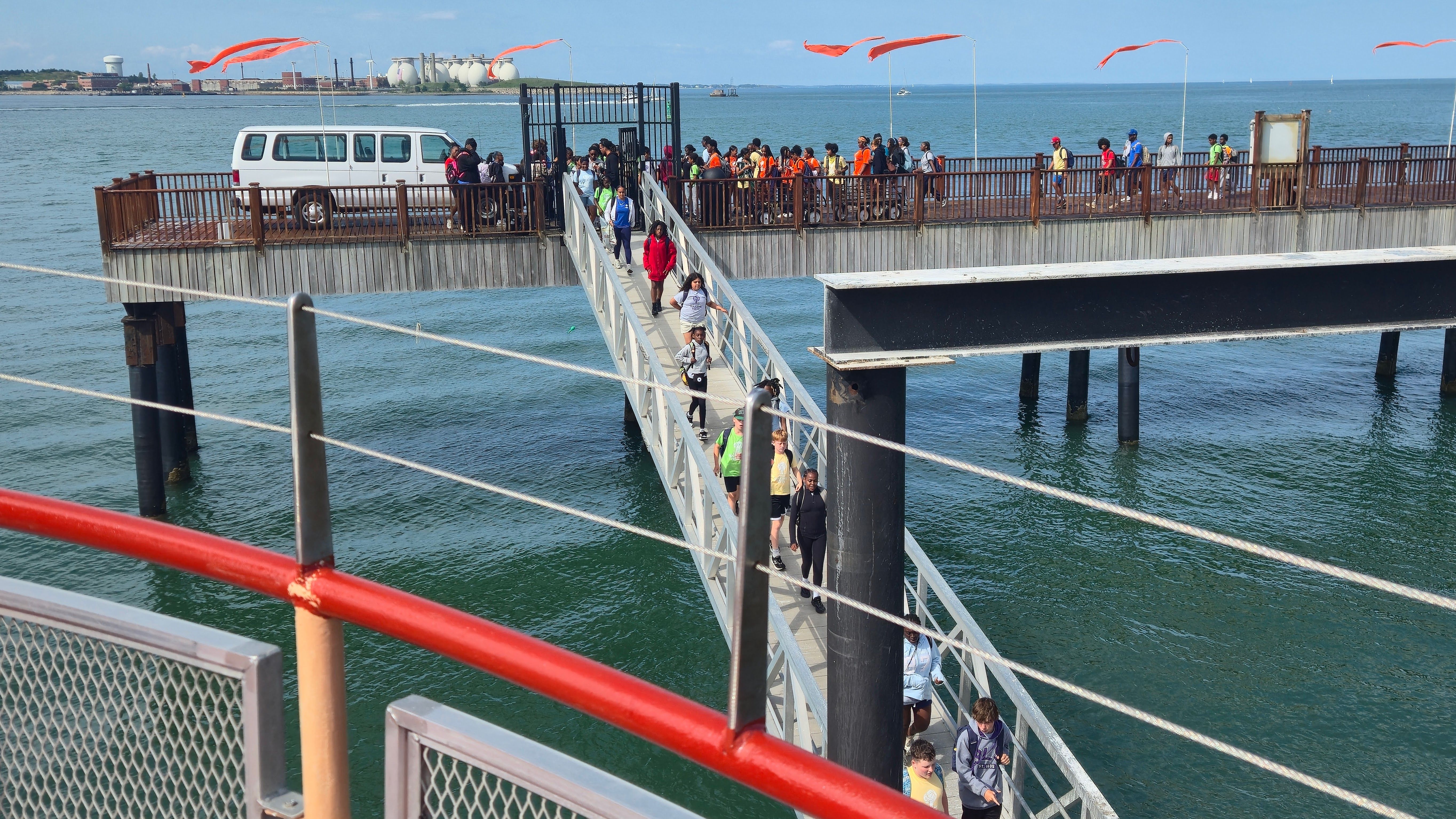 A crowded walkway extends over blue water with railings; many people in colorful clothes walk along the pier, a white van is parked on the platform, and orange banners flutter overhead.