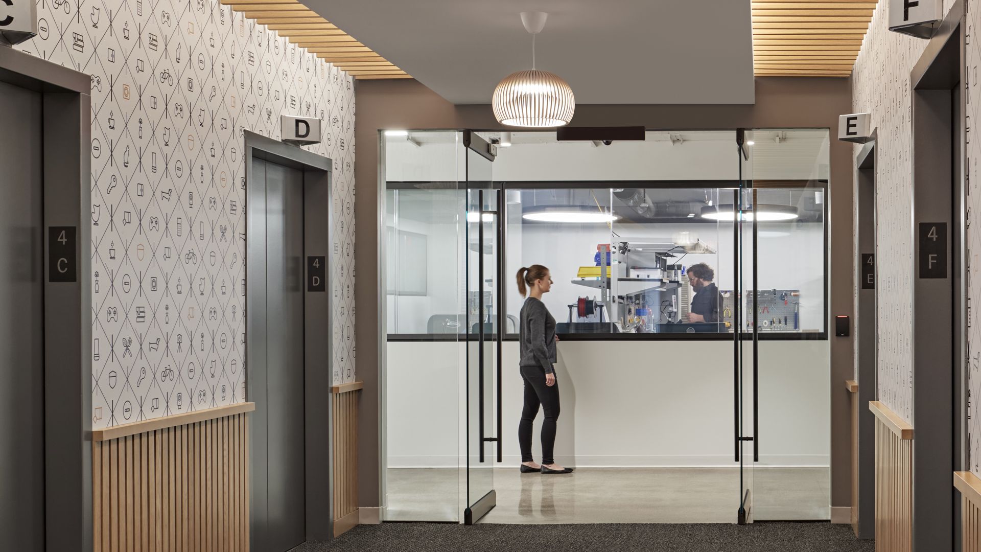 Past the elevators and entrance, a woman looks through the window into SimpliSafe's dry lab where an employee is working.