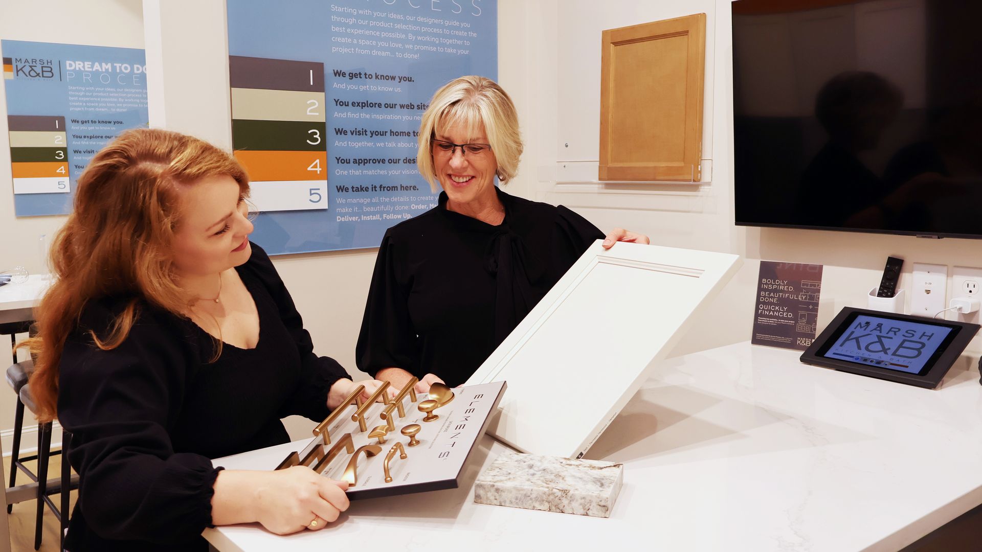 Two women in a meeting area. One is holding a kitchen cabinet door, the other is holding samples of cabinet handles.