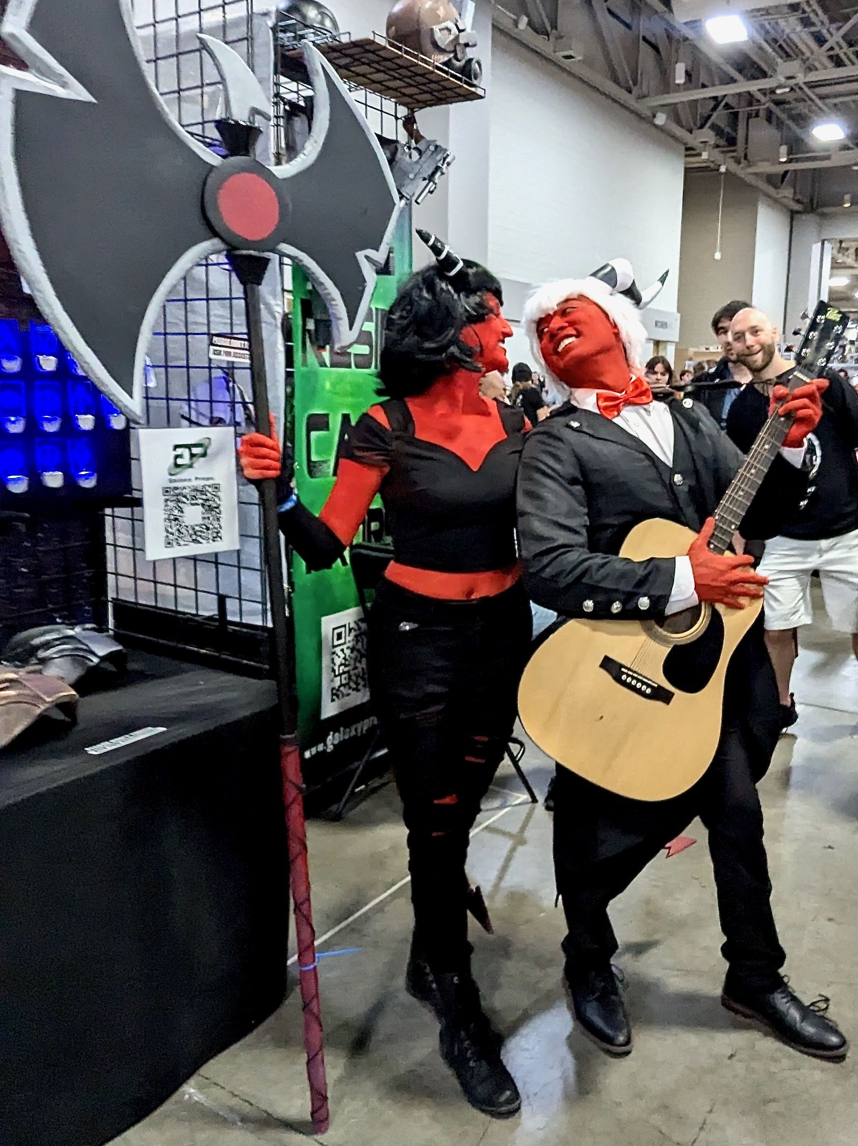 People costumed as Millie and Moxxie from Helluva Boss pose lovingly with a large battleaxe and a guitar.