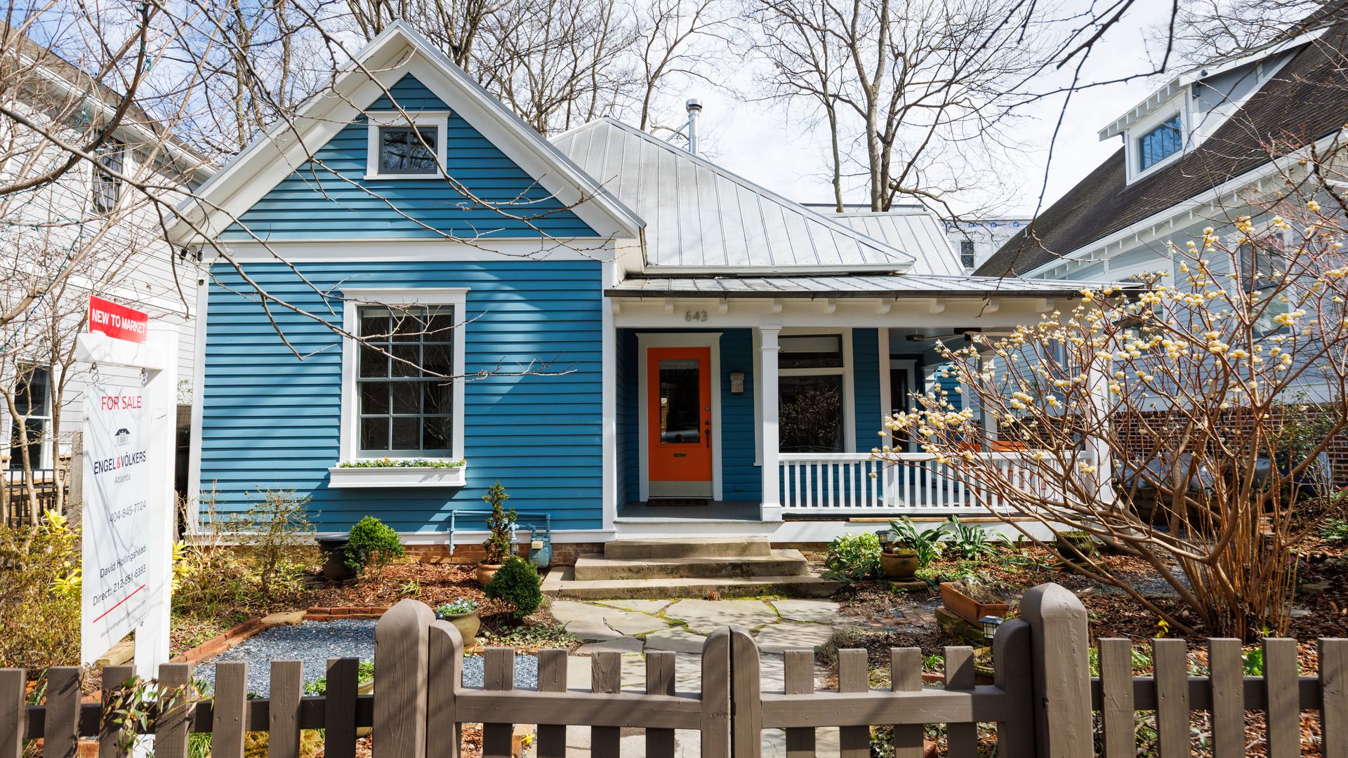 A blue house sits behind a fence with a for-sale sign in the yard.