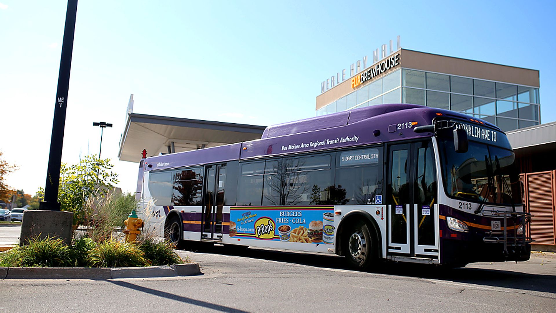 Purple and white Des Moines Area Regional Transit Authority bus 2113 parked beside a modern glass building with a "FLEX BREWHOUSE" sign; side ad shows burgers, fries, and a cola.