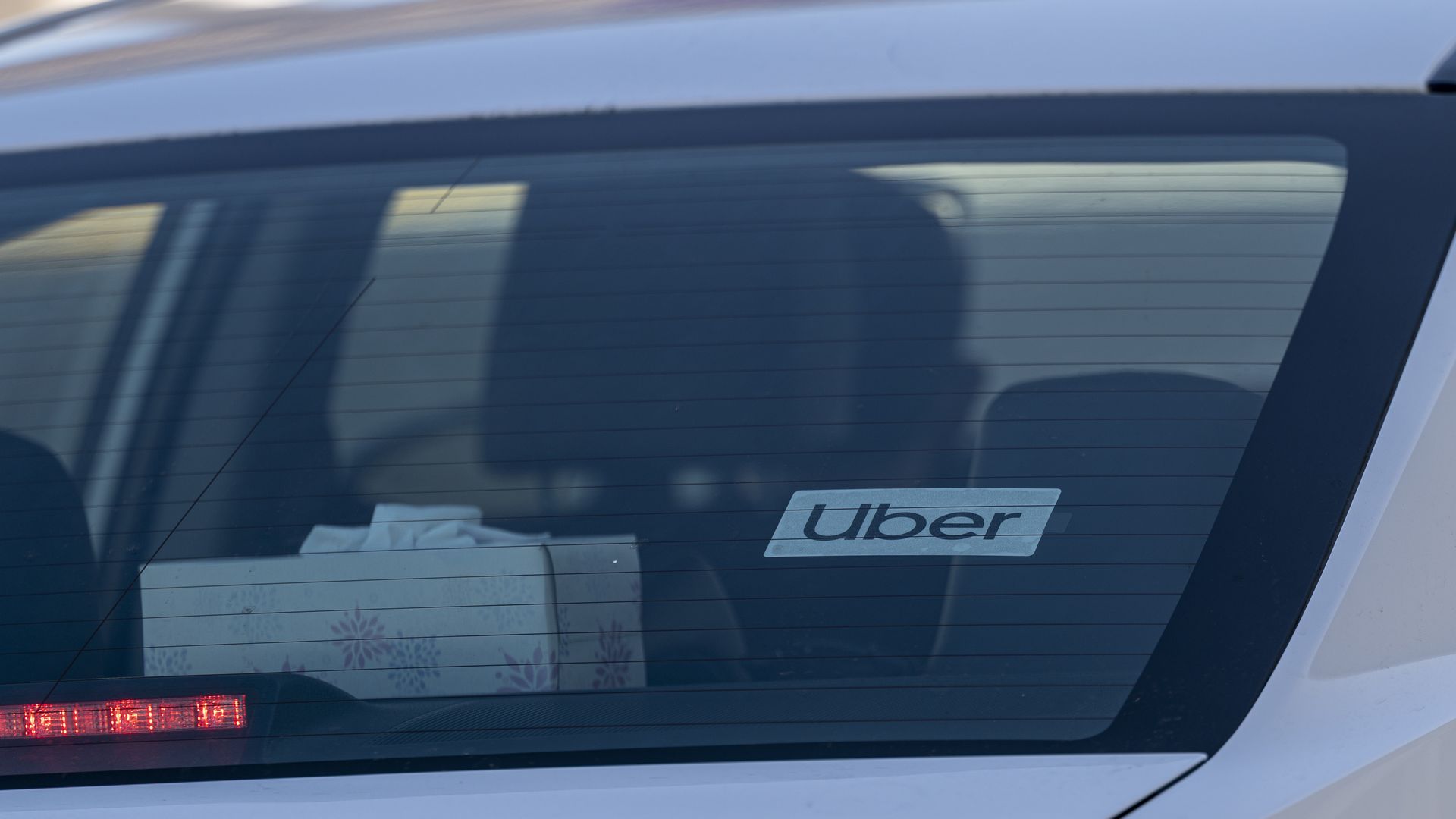 A white sticker that says Uber in black appears on the back windshield of a white car at an airport.