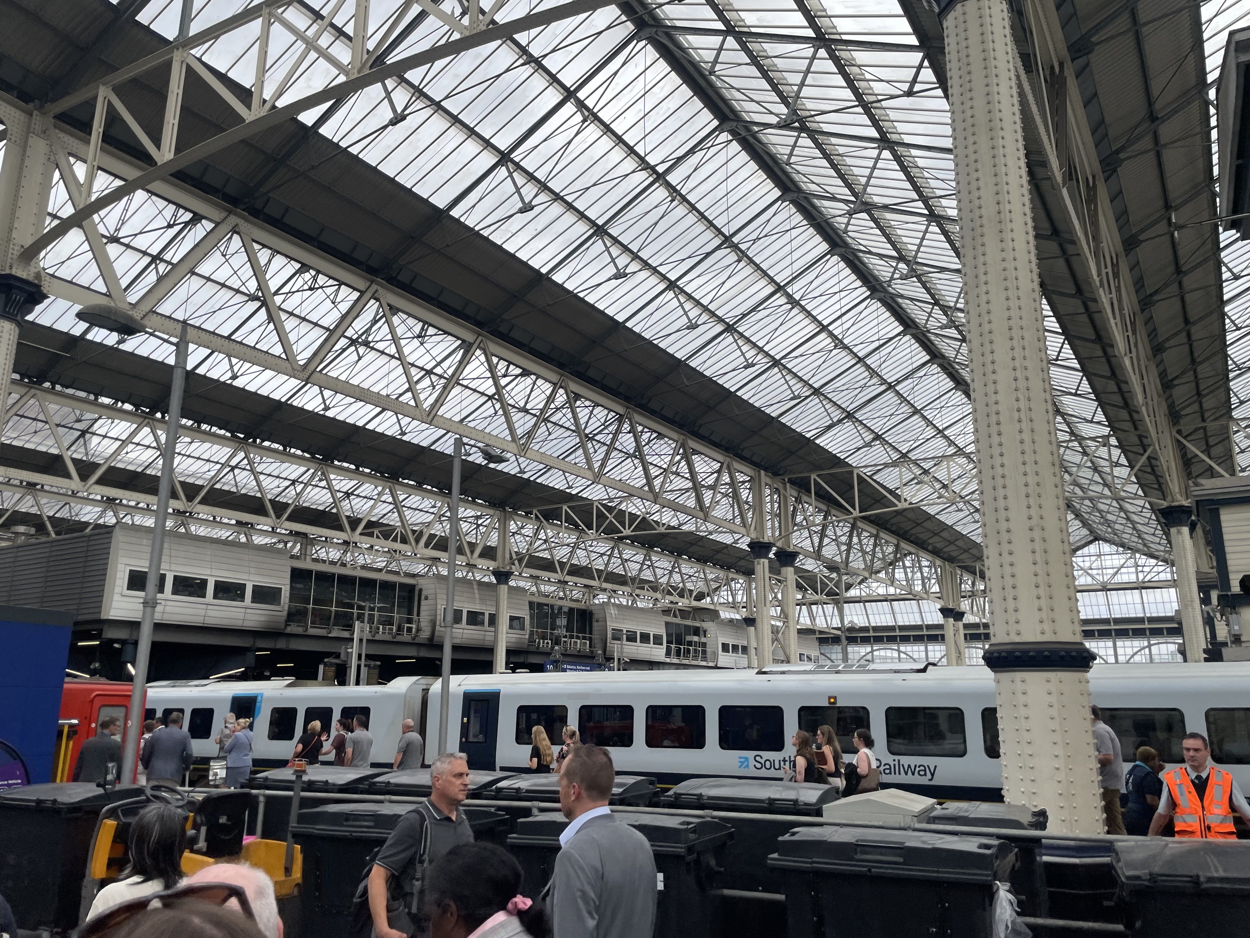 Interior of a large railway station featuring a glass and steel vaulted roof, white riveted columns, and a platform with a white train and passengers waiting.