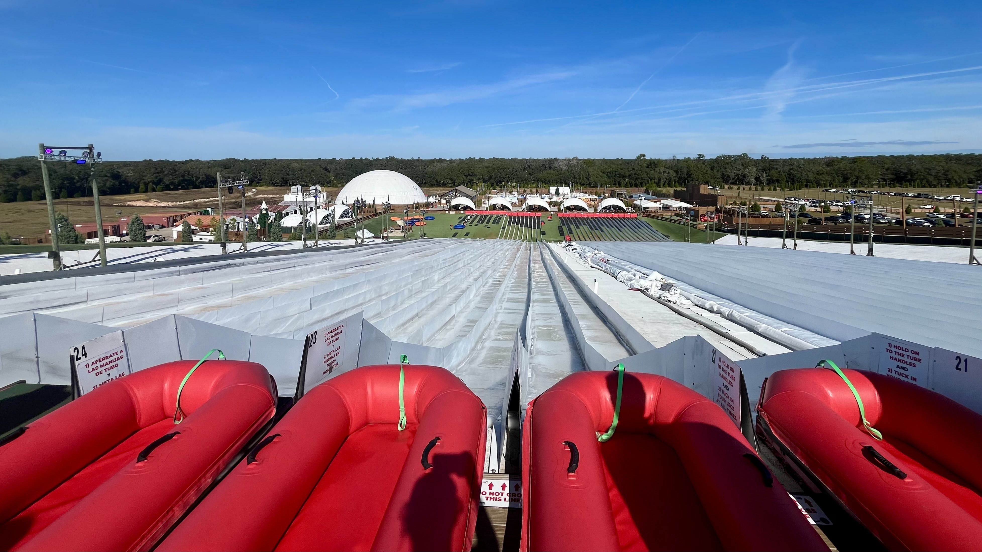 View from top of a giant white multi-lane slide with red inflatable tubes, set in an open area with green grass, white tents, a dome structure, and blue sky.