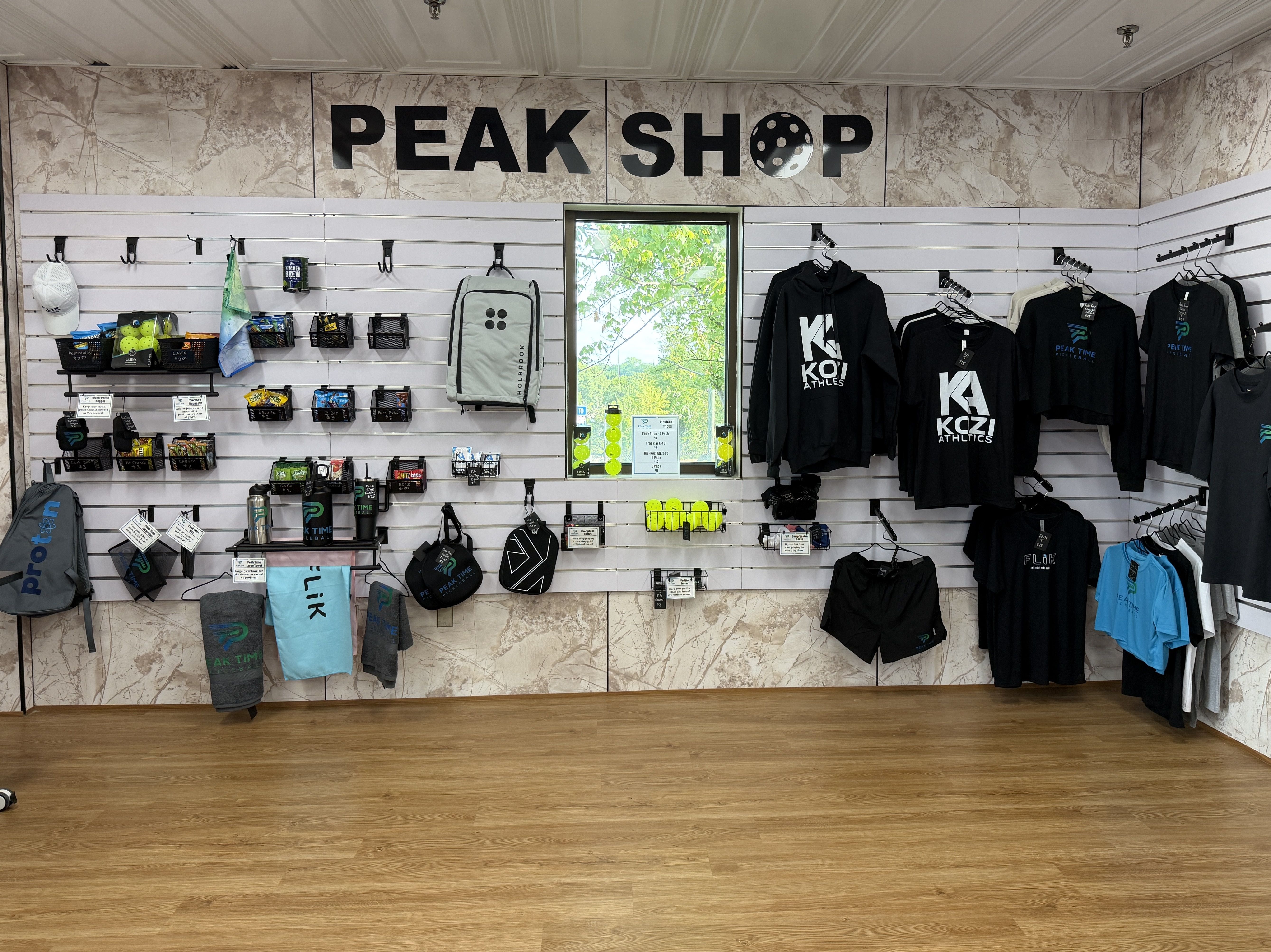 Interior of Peak Shop with marble walls and a large black sign reading "PEAK SHOP", white slat displays holding clothing, bags, and gear; a window shows green trees outside.