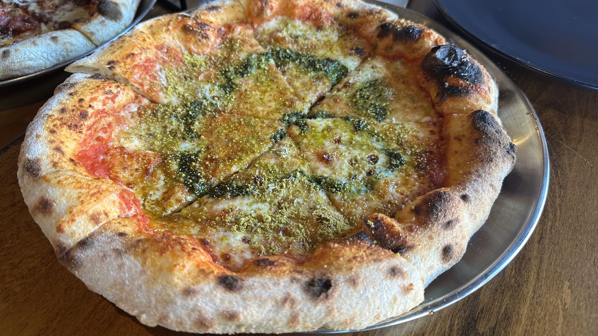 Rustic pizza on a metal tray; the foreground pie has bubbly crust, melted cheese, red tomato sauce and a green pesto-like topping, with charred bubbles along the edge.