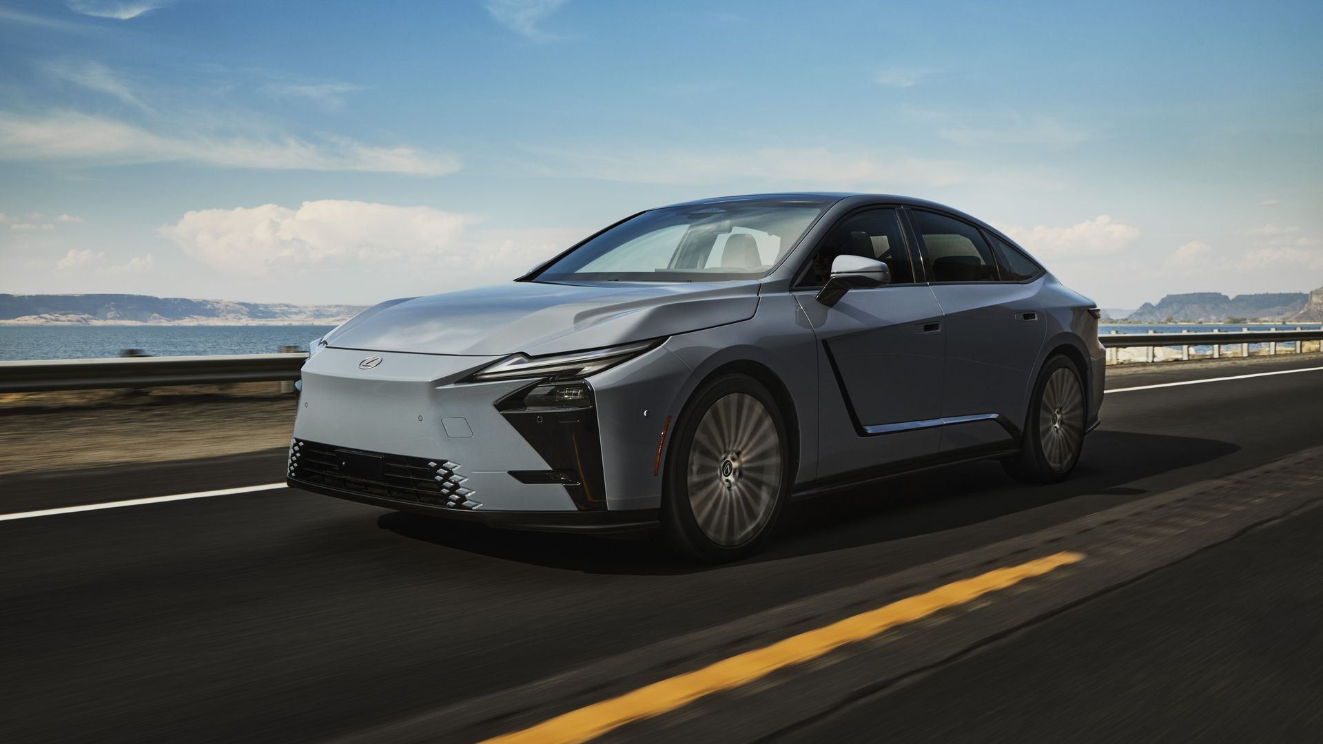 Silvery-blue Lexus ES 350 electric sedan in motion on a coastal highway by the sea, with a guardrail, blue sky and clouds, distant cliffs in the background; front three-quarter view.